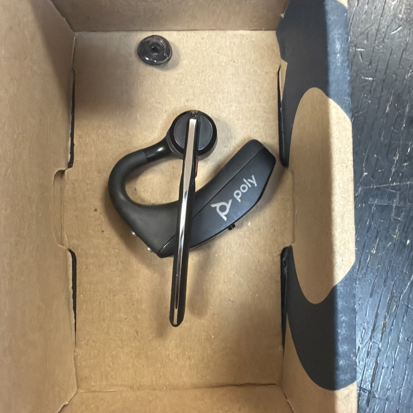 Black wireless earpiece with a silver microphone arm and the brand name "poly" printed on the side, resting inside a plain cardboard box.