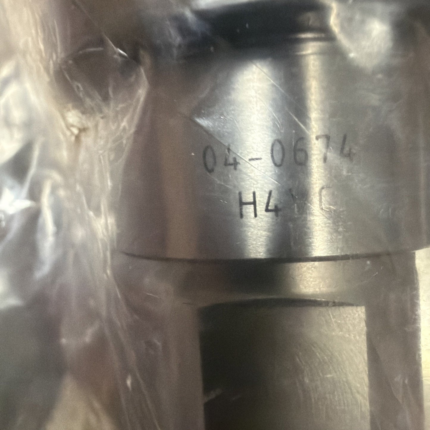 Close-up of a metallic cylindrical component partially wrapped in clear plastic, displaying engraved text "04-0674" and "H41 C" on its smooth surface.
