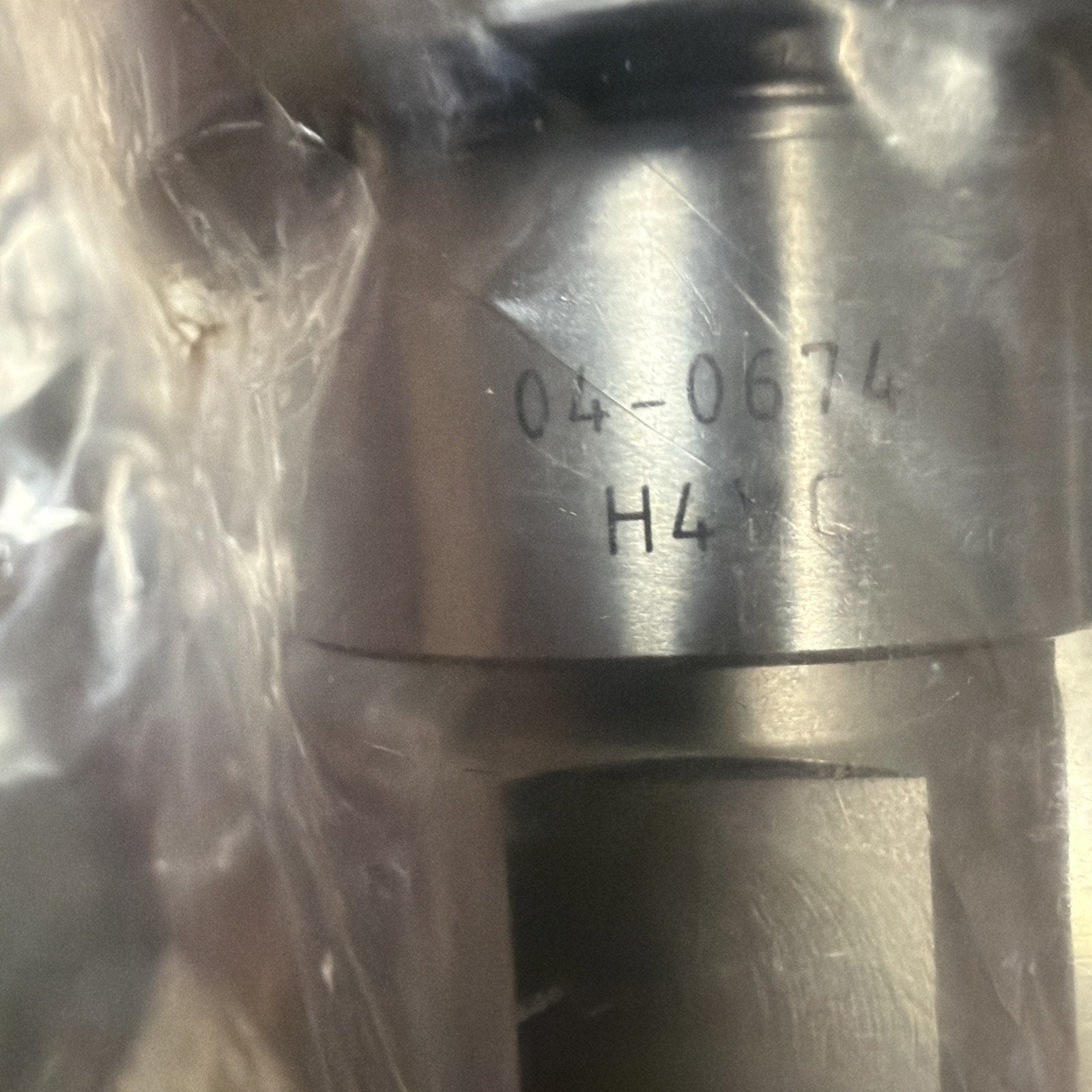 Close-up of a metallic cylindrical component partially wrapped in clear plastic, displaying engraved text "04-0674" and "H41 C" on its smooth surface.