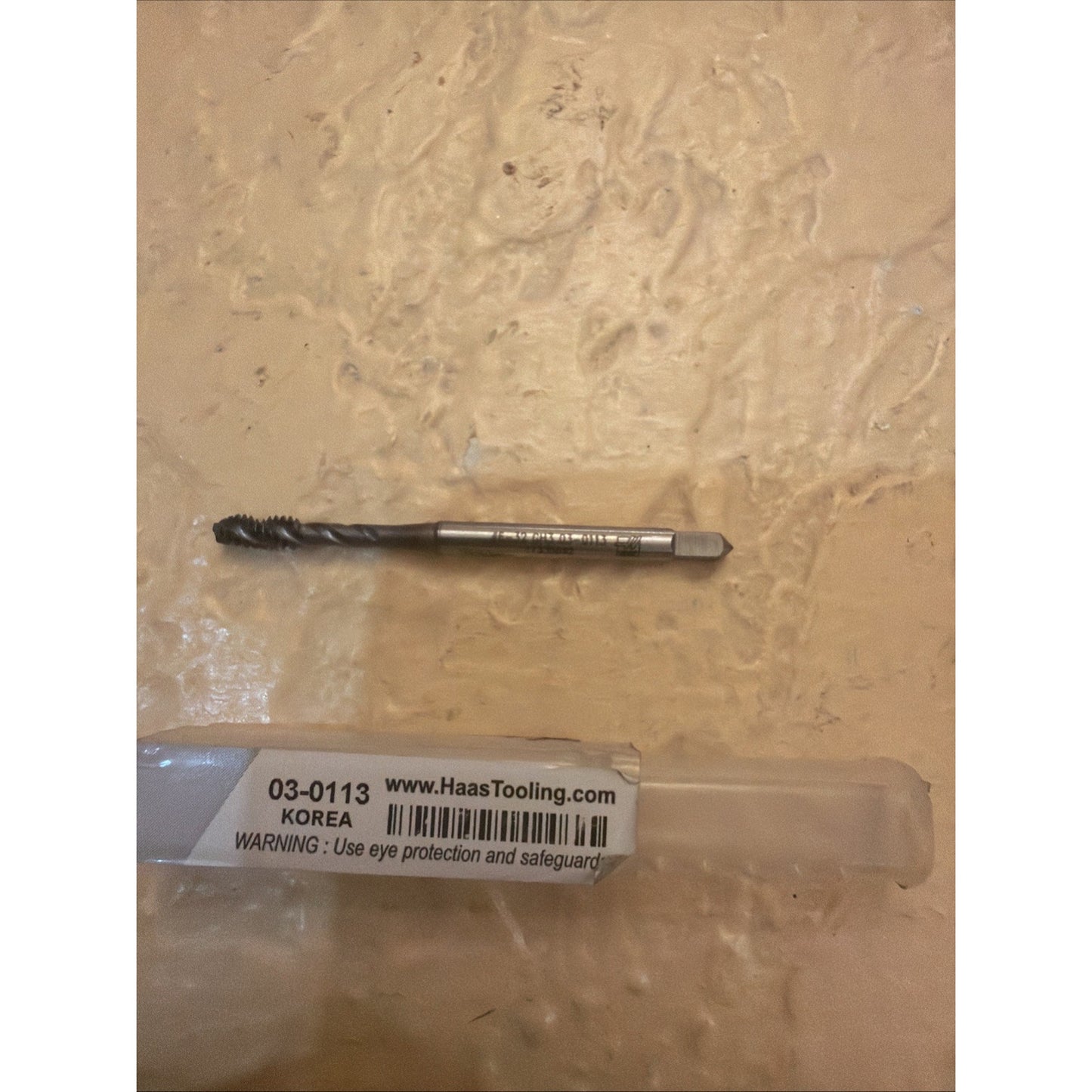 A single metal threading tap with helical flutes and a square drive tip resting on a textured beige surface above a transparent plastic case labeled with safety warnings and a web address.