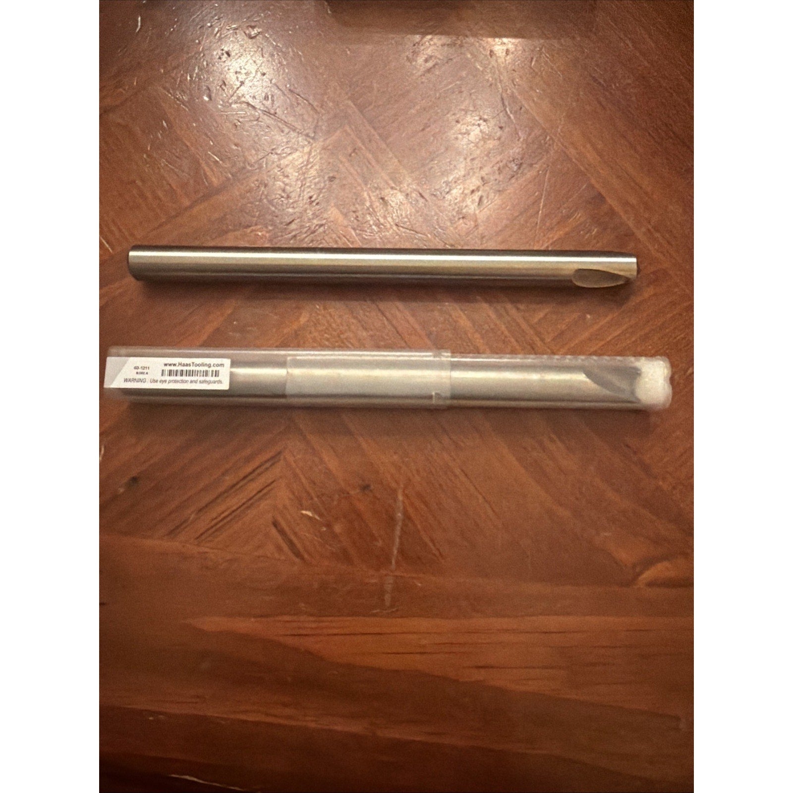 Metal drill bit with a smooth cylindrical shaft and a single cutting edge, displayed alongside a clear plastic protective tube with a white label and barcode, resting on a wooden surface.