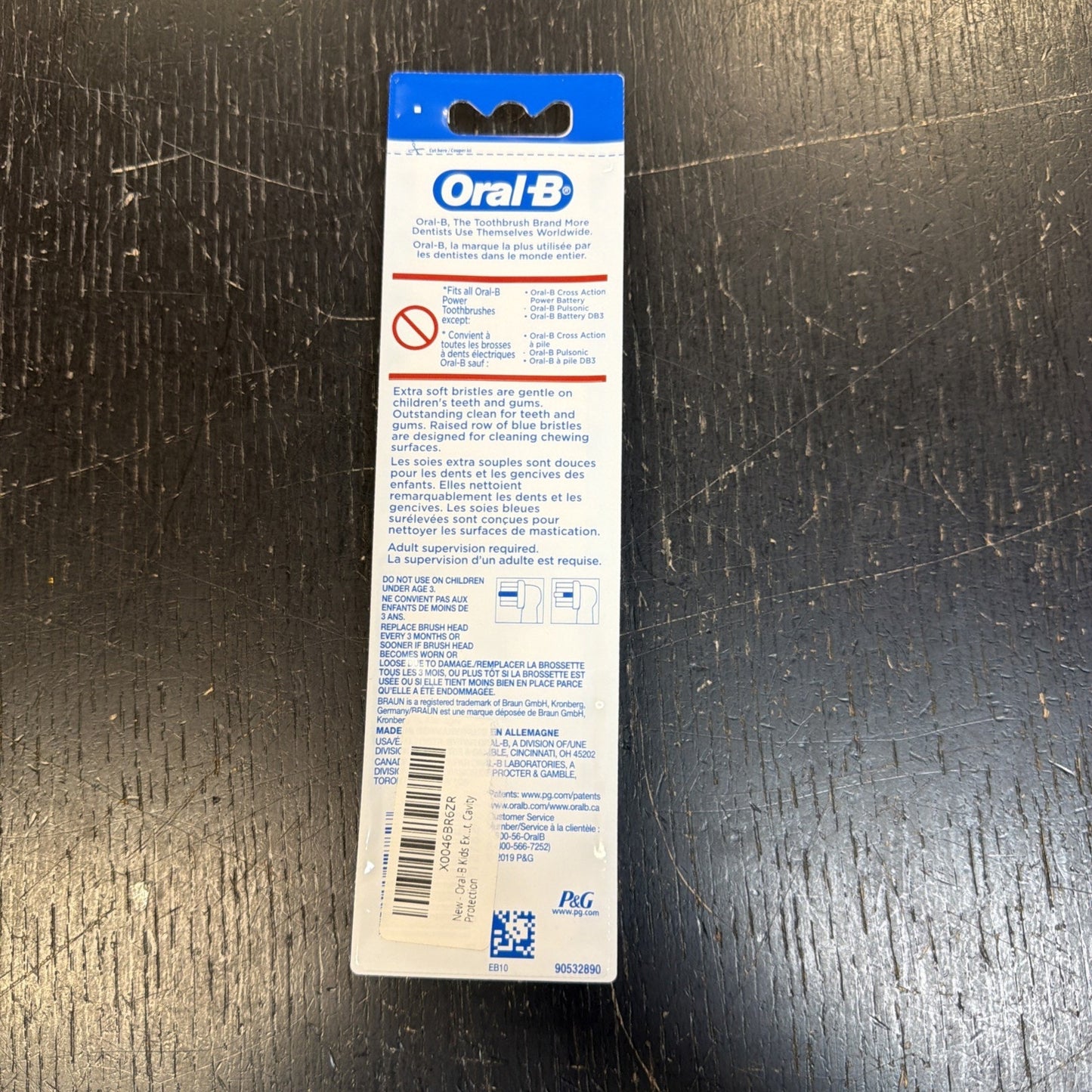 Back view of an Oral-B toothbrush package with detailed usage instructions, warnings, product features, barcode, and company information on a white and blue label against a dark textured background.