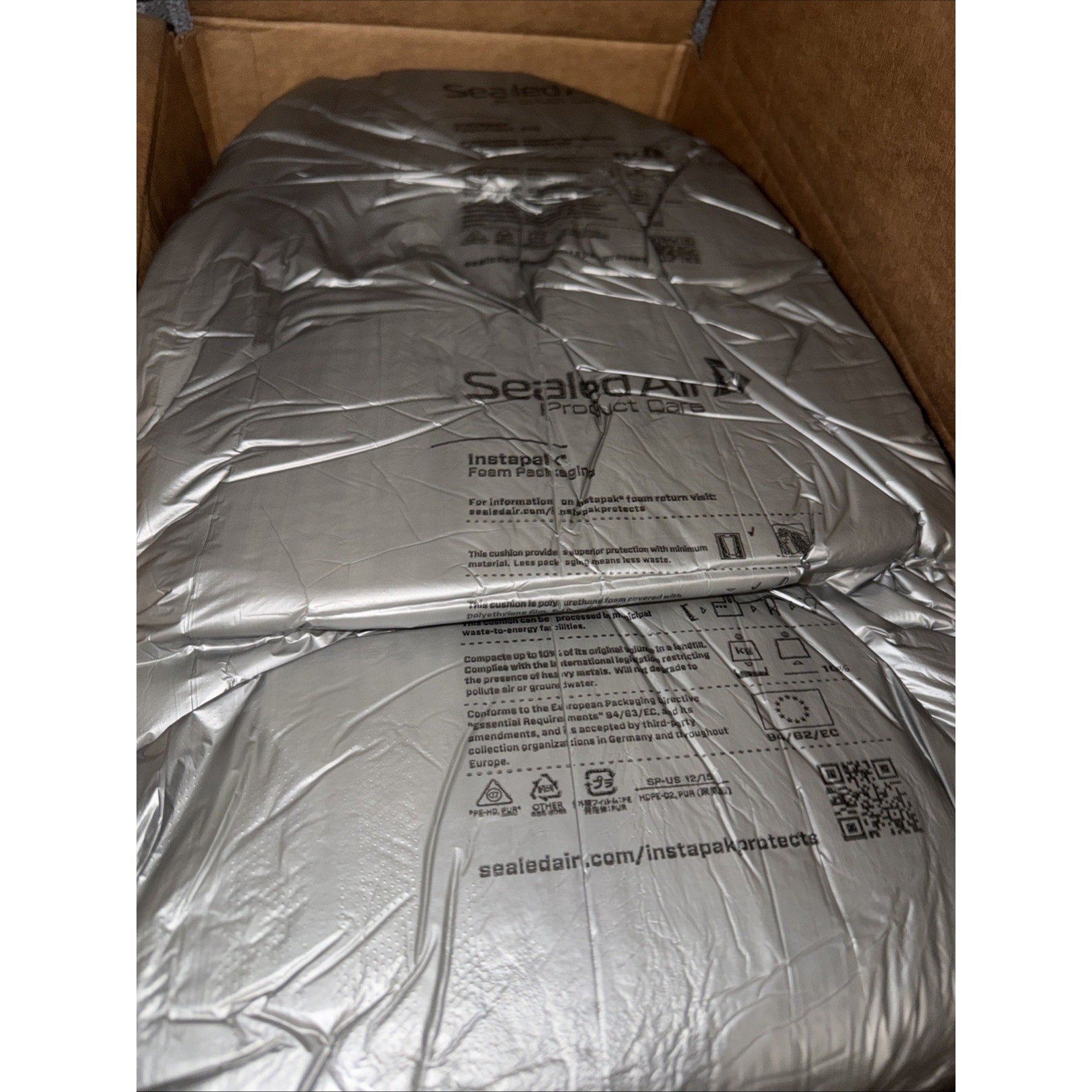 Silver inflatable air cushion packaging labeled "Sealed Air Instapak Foam Pads" inside a brown cardboard box, with printed instructions and a QR code visible on the surface.