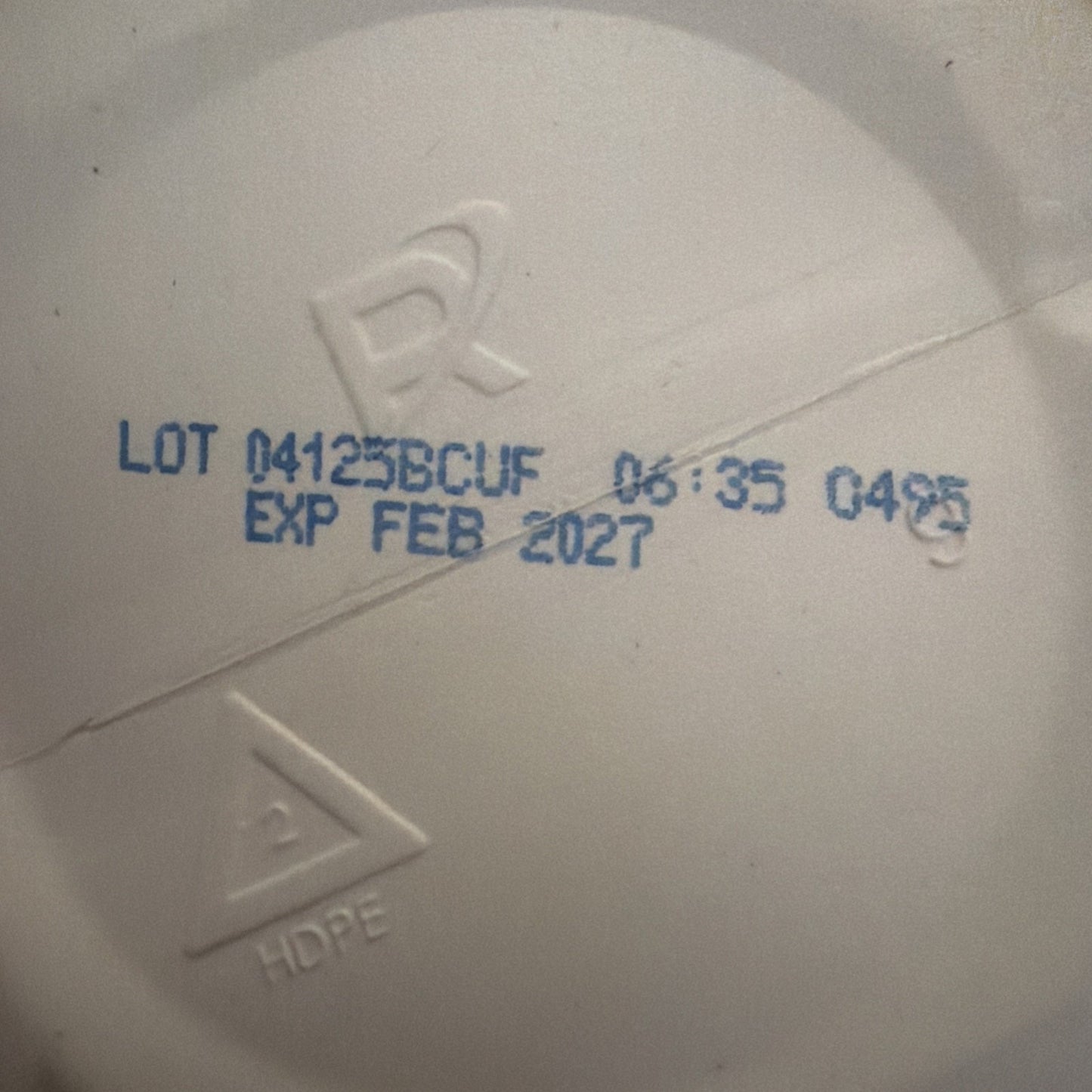 Close-up of a white plastic surface with blue printed lot number "04125BCUF," time stamp "06:35," code "0425," and expiration date "FEB 2027," showing HDPE recycling symbol.