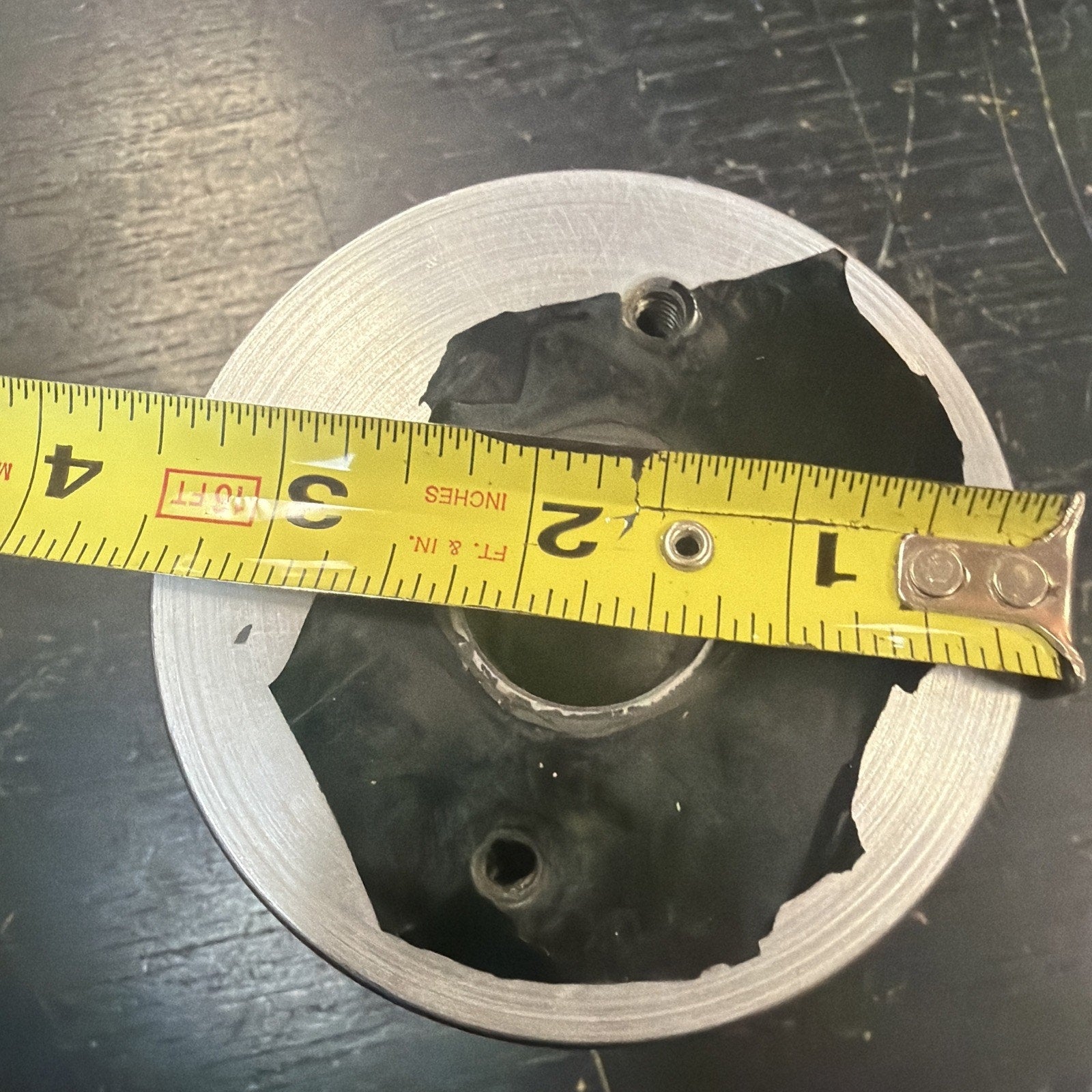 Close-up of a circular metal object with a black center piece and two threaded holes, with a yellow measuring tape stretched across showing approximately 3 inches.