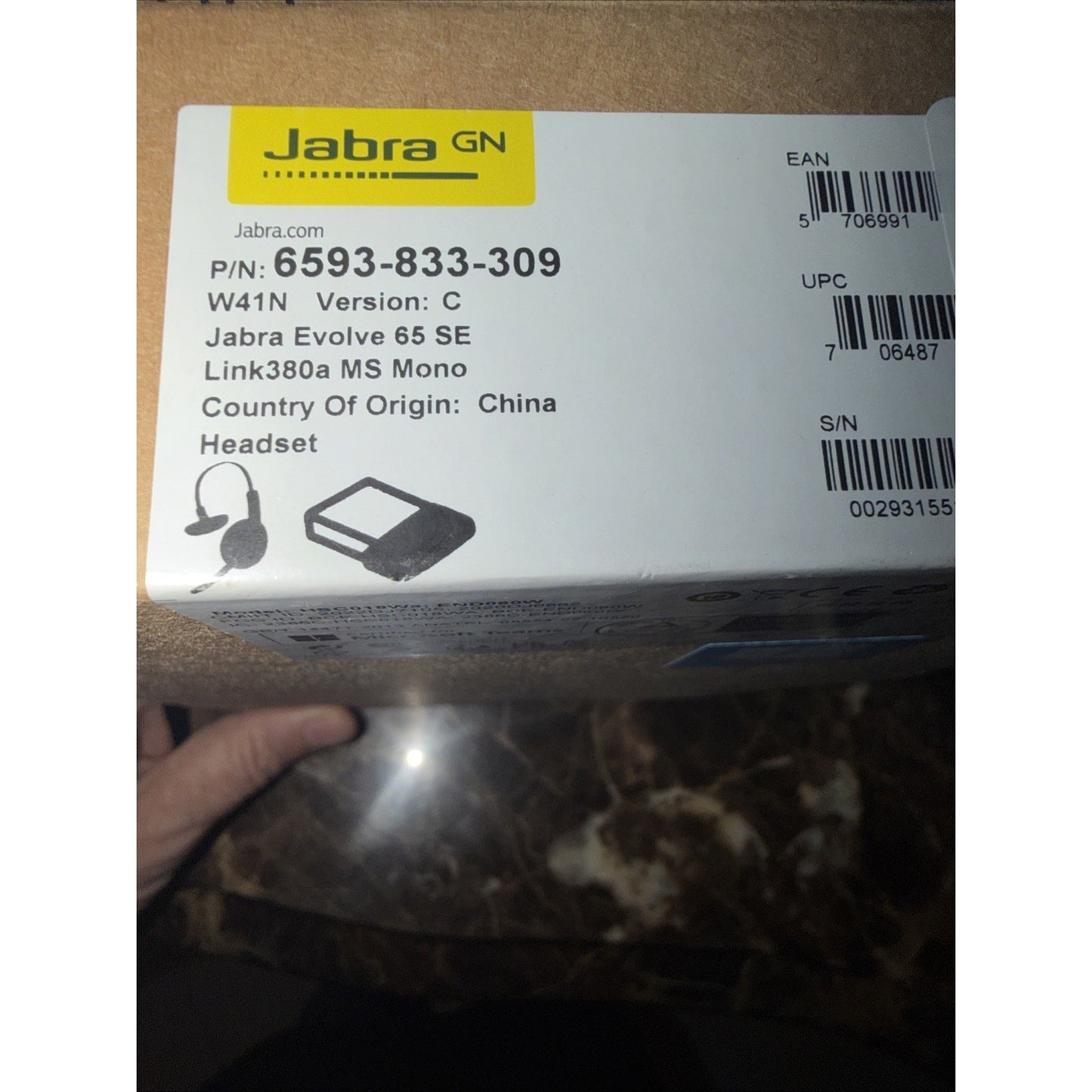Close-up of a white product label on a brown cardboard box showing Jabra branding, product number, model details for a Jabra Evolve 65 SE headset, part illustrations, and manufacturing origin.