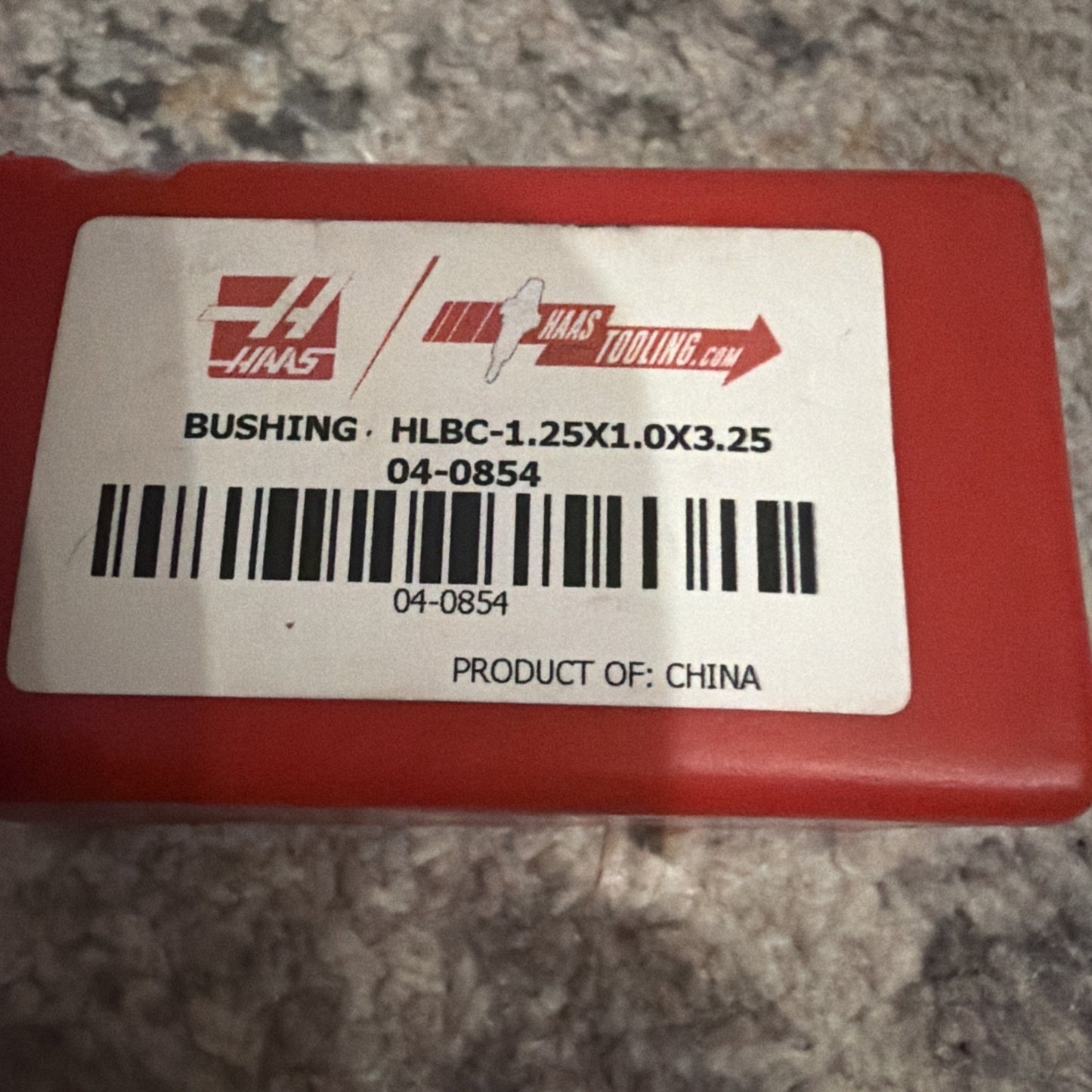 Red plastic container with a white label displaying the Haas logo, product code, barcode, and text indicating the item is a bushing made in China, sitting on a textured surface.