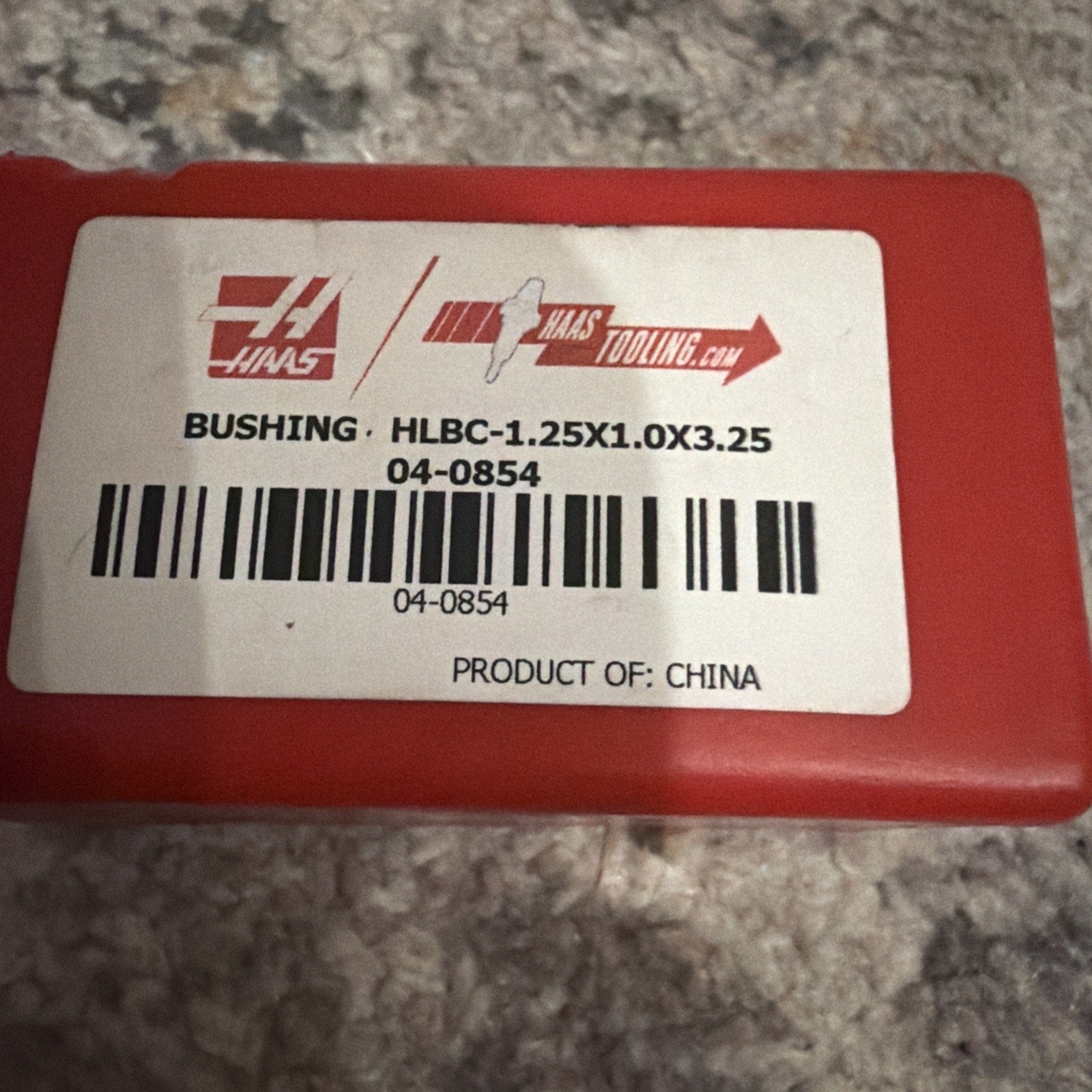 Red plastic container with a white label displaying the Haas logo, product code, barcode, and text indicating the item is a bushing made in China, sitting on a textured surface.