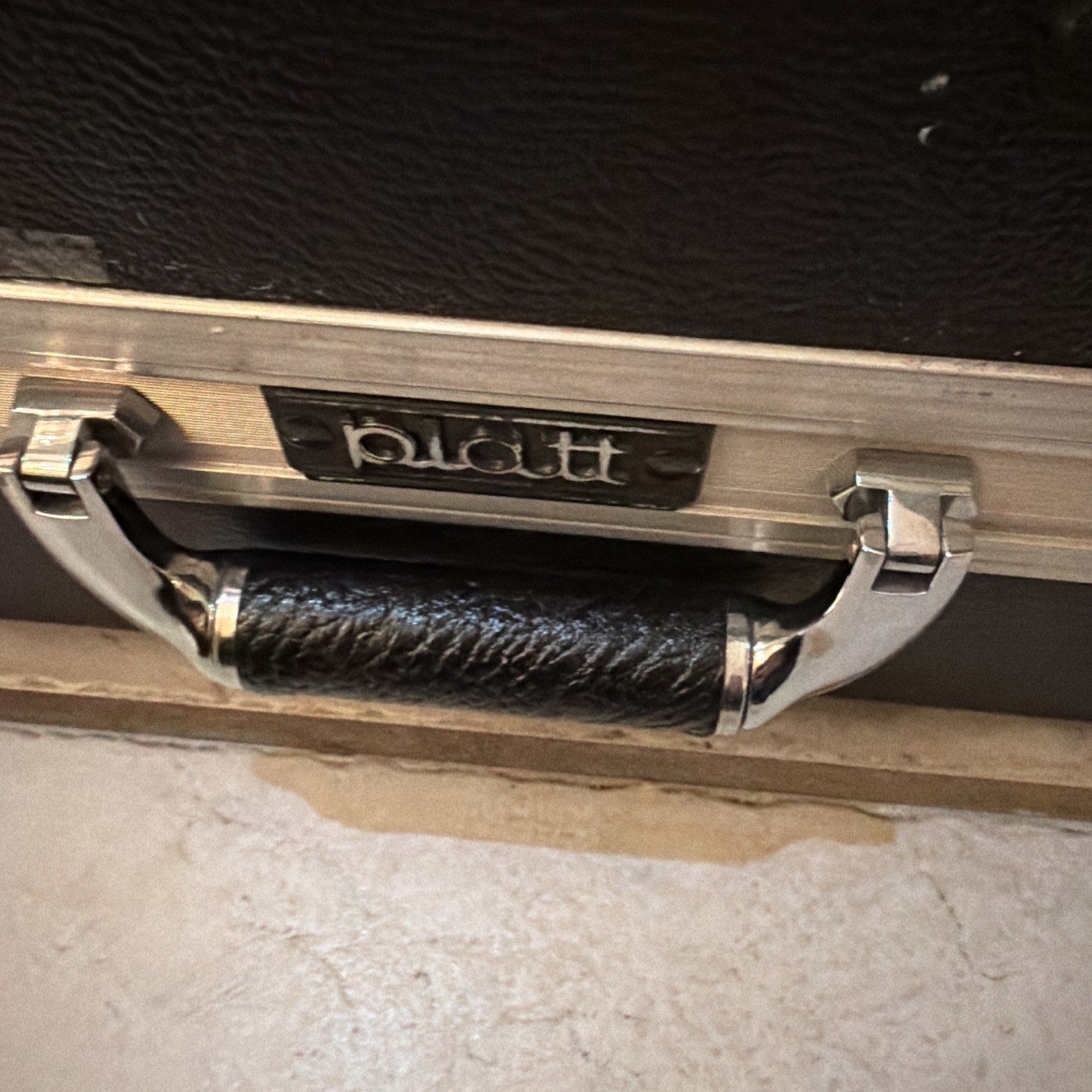 Close-up of a black textured handle with silver metal brackets attached to a black case, featuring a small metal nameplate with the word "platt.