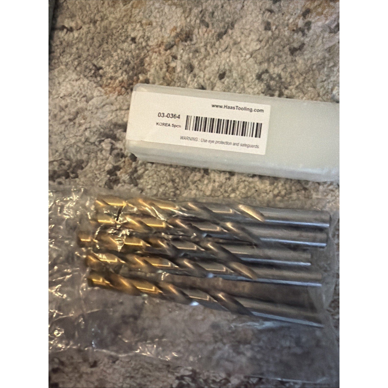 Five metal twist drill bits with silver and gold-colored fluted sections, sealed in a clear plastic bag, placed on a textured surface next to a white storage box with a barcode label.
