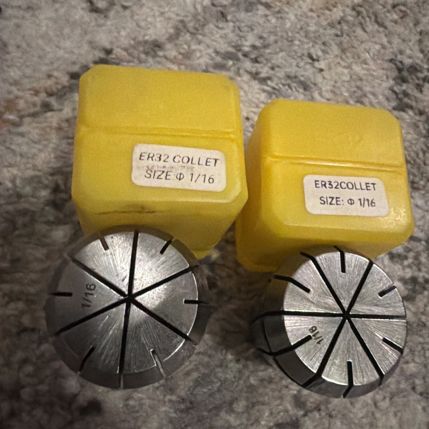 Two metal ER32 collets with engraved size 1/16, placed in front of corresponding yellow plastic storage cases labeled with size and type, resting on a textured surface.