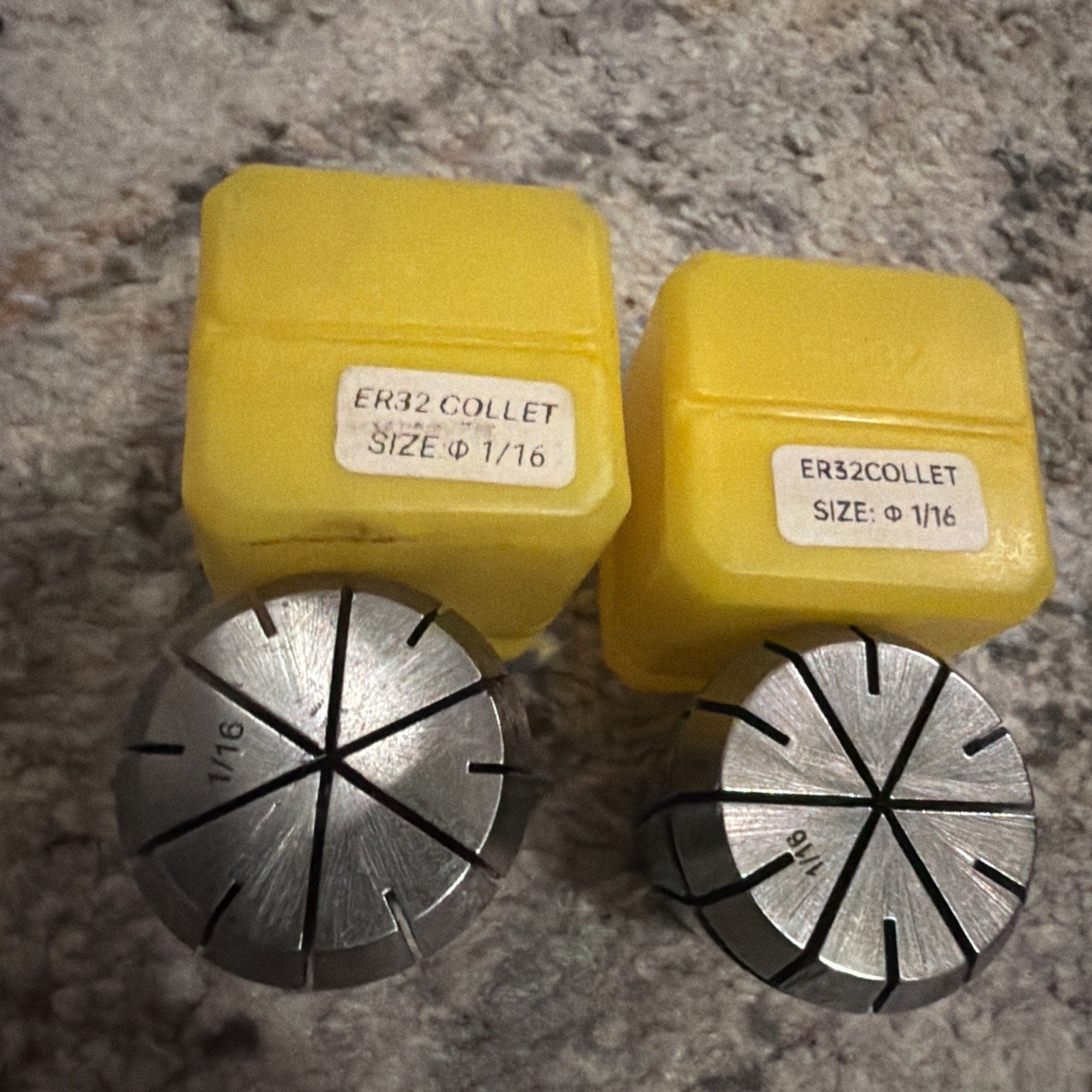 Two metal ER32 collets with engraved size 1/16, placed in front of corresponding yellow plastic storage cases labeled with size and type, resting on a textured surface.