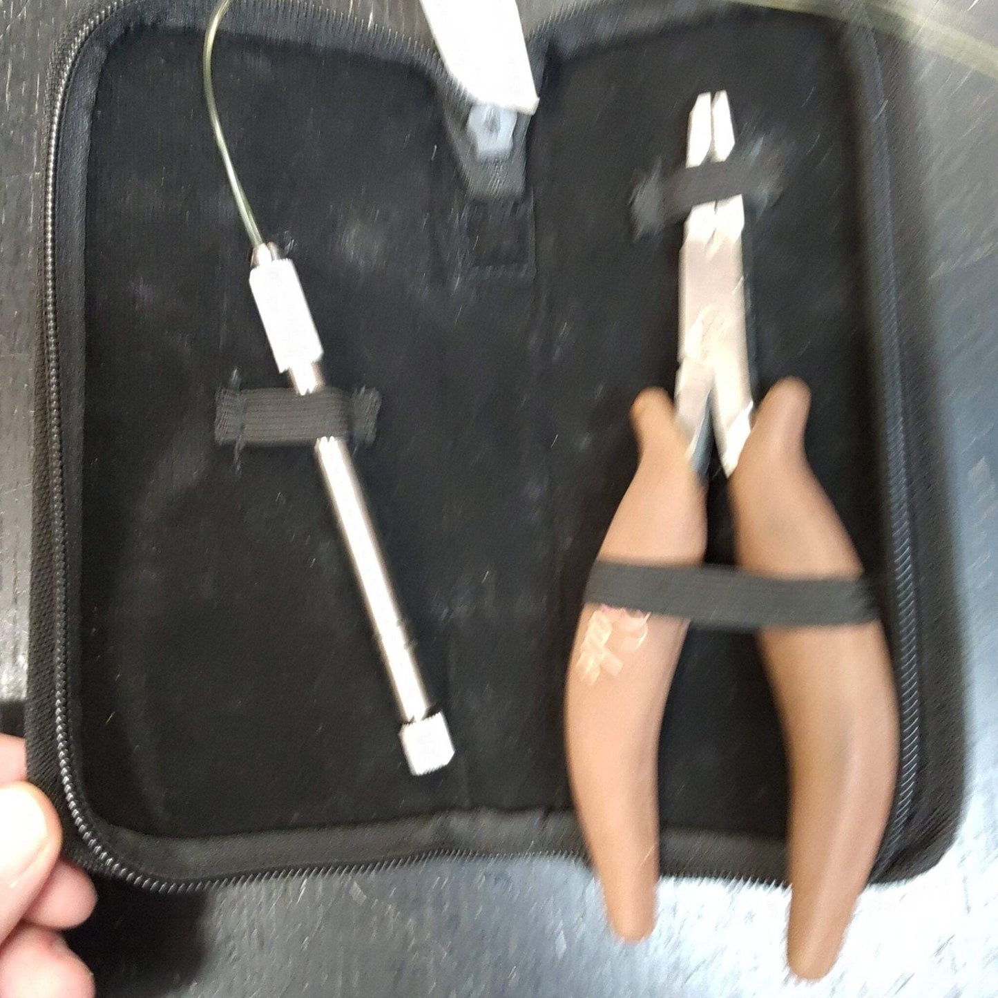 Black zippered case holding a pair of needle-nose pliers with brown handles and a slim metal rod tool secured in elastic straps against a soft black interior lining.