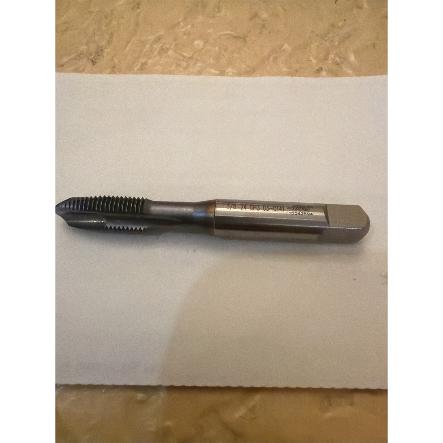 Single metal threading tap with a polished shank and sharp cutting threads, lying horizontally on a white sheet over a beige textured surface.