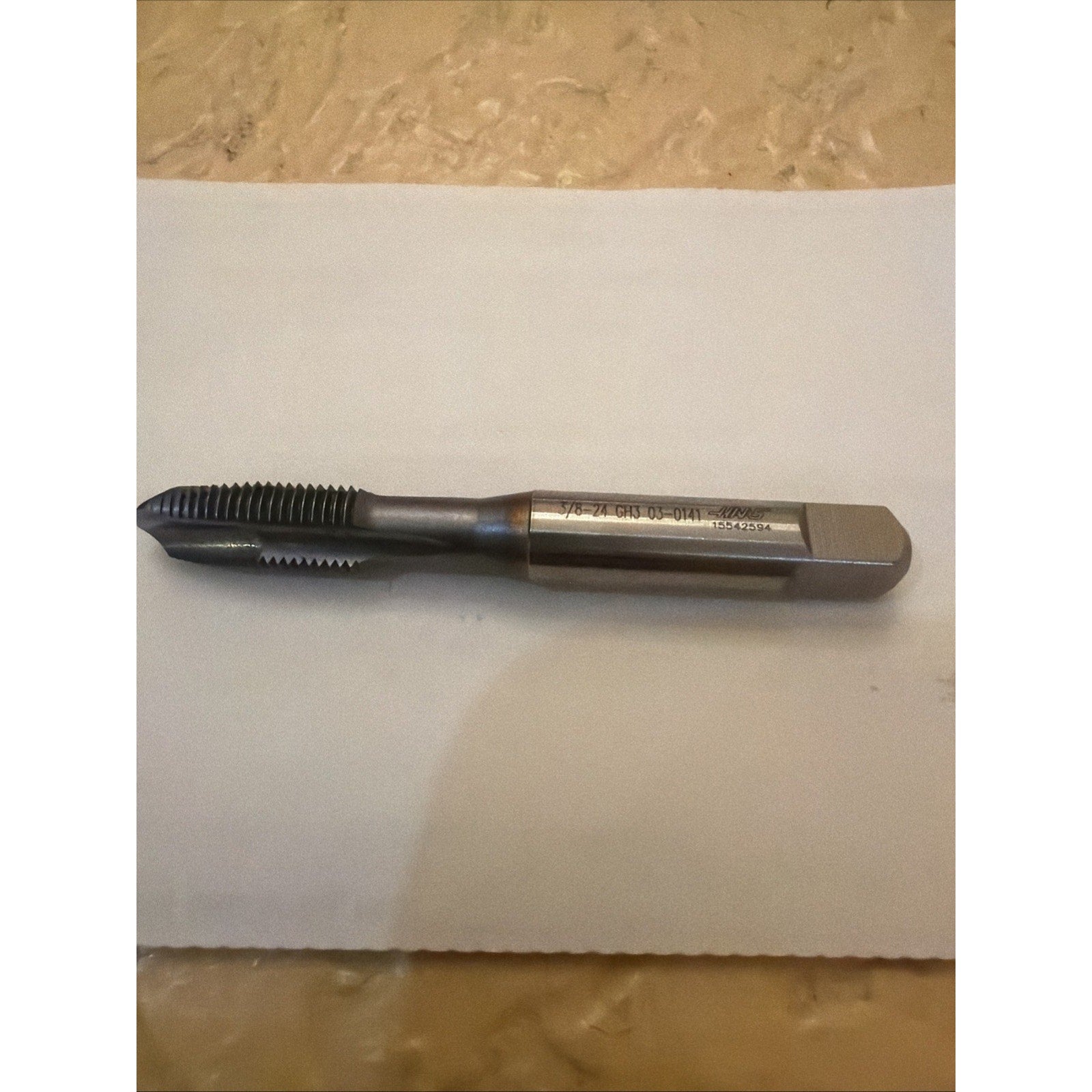 Single metal threading tap with a polished shank and sharp cutting threads, lying horizontally on a white sheet over a beige textured surface.