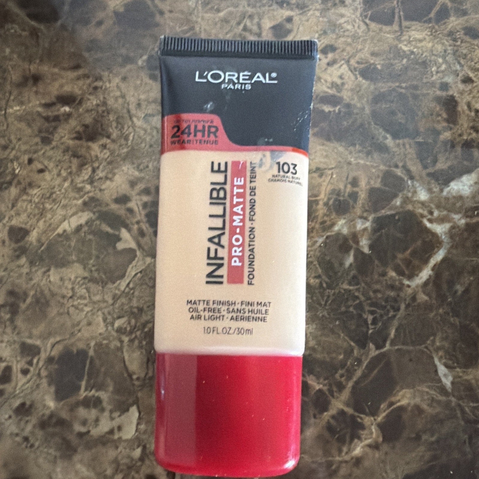 Tube of L'Oréal Paris Infallible Pro-Matte liquid foundation in shade 103 True Beige with a black cap and red base, placed on a dark marbled surface.