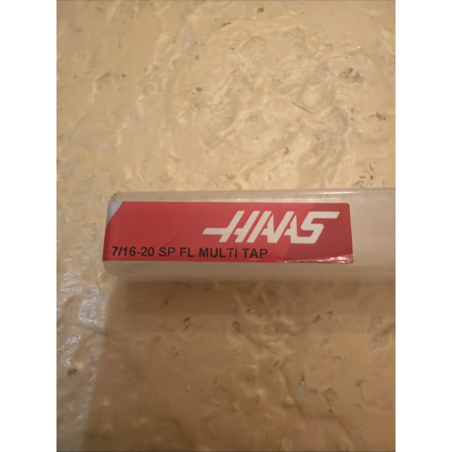 Clear rectangular plastic storage container with a red label displaying white text including "7/16-20 SP FL MULTI TAP" and a stylized white "HAAS" logo on a textured beige surface.