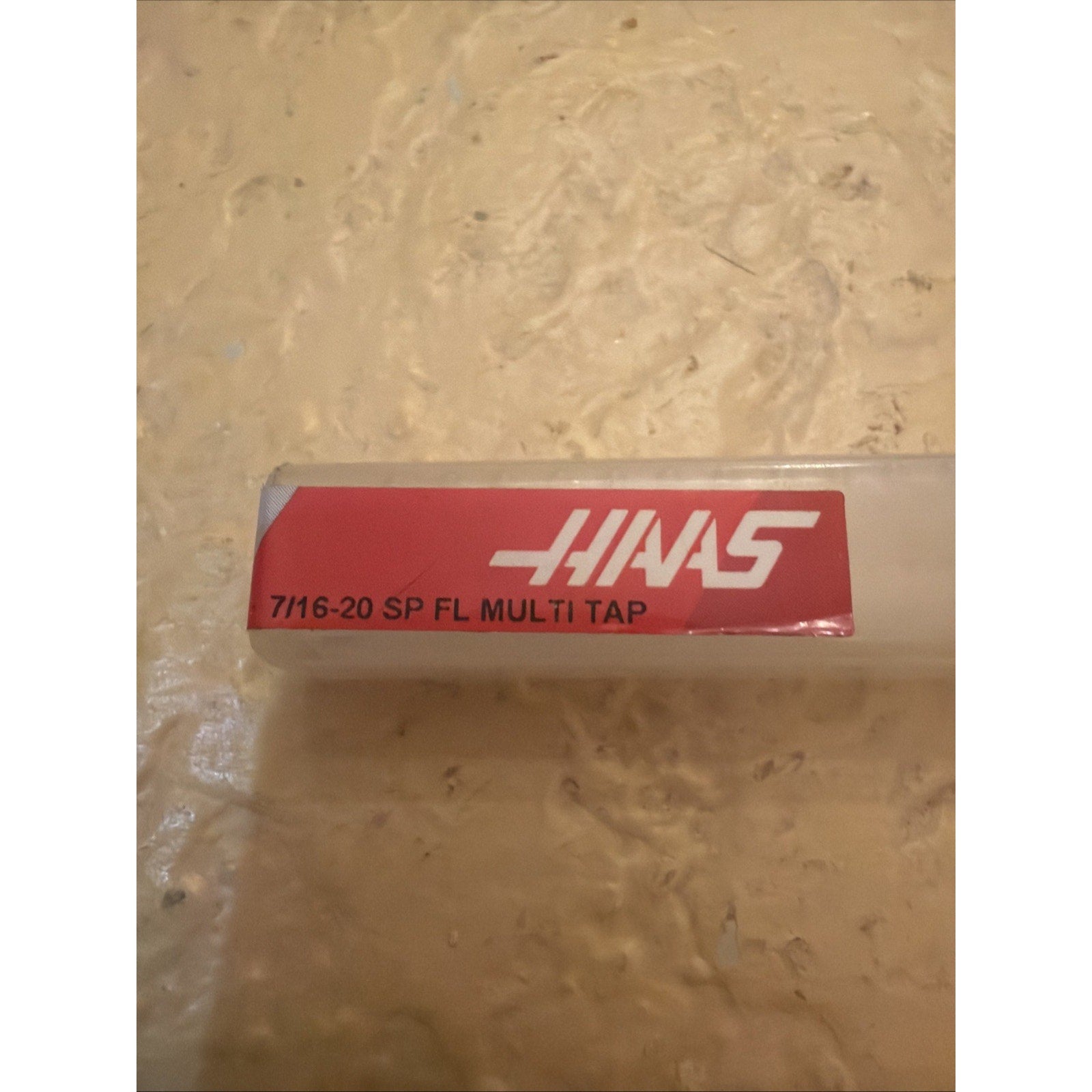 Clear rectangular plastic storage container with a red label displaying white text including "7/16-20 SP FL MULTI TAP" and a stylized white "HAAS" logo on a textured beige surface.