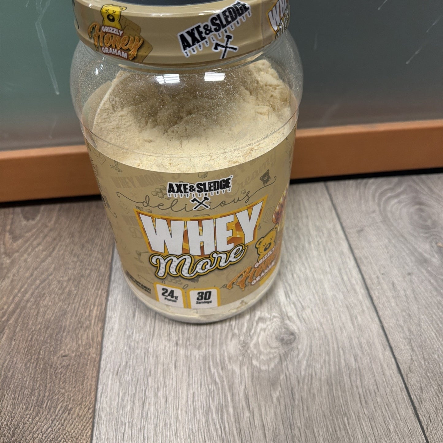 Open container of Axe & Sledge Whey More protein powder in peanut butter flavor, showing light beige powder inside and label indicating 24 grams of protein and 30 servings.