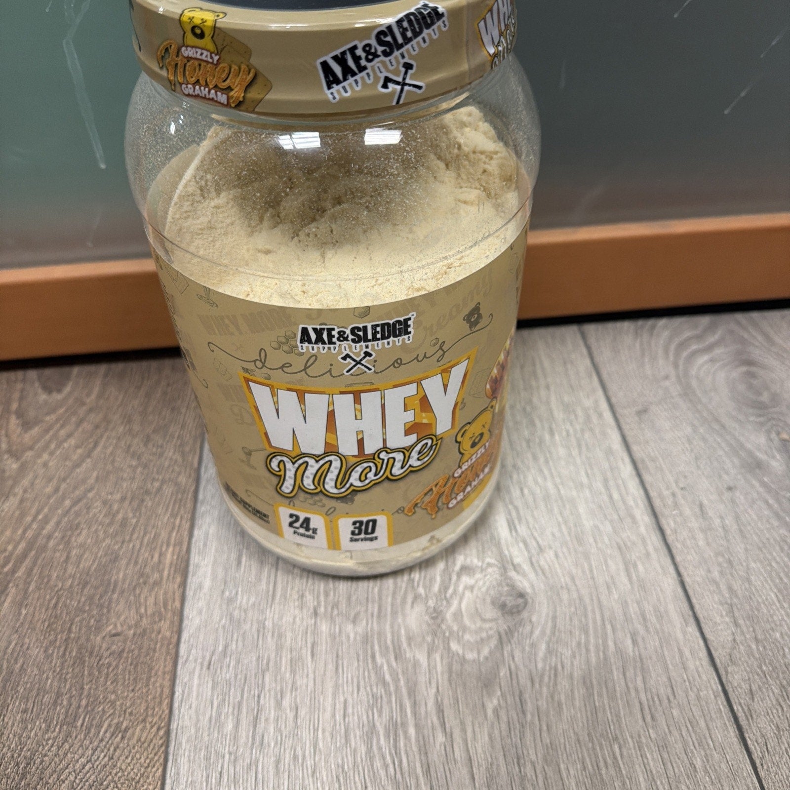 Open container of Axe & Sledge Whey More protein powder in peanut butter flavor, showing light beige powder inside and label indicating 24 grams of protein and 30 servings.