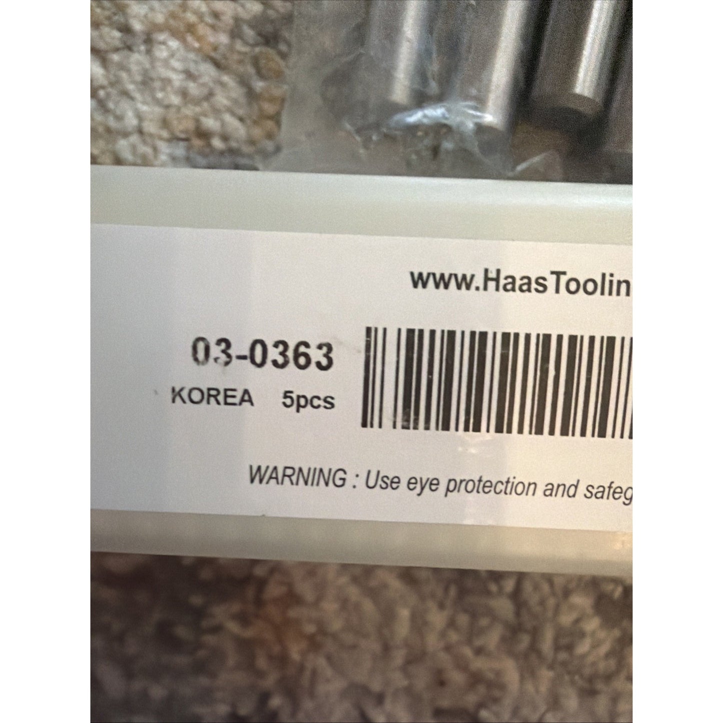 Close-up of a white label on clear plastic packaging showing a barcode, product number, origin marked as Korea, quantity of 5 pieces, and a safety warning about eye protection, placed on a beige textured surface.