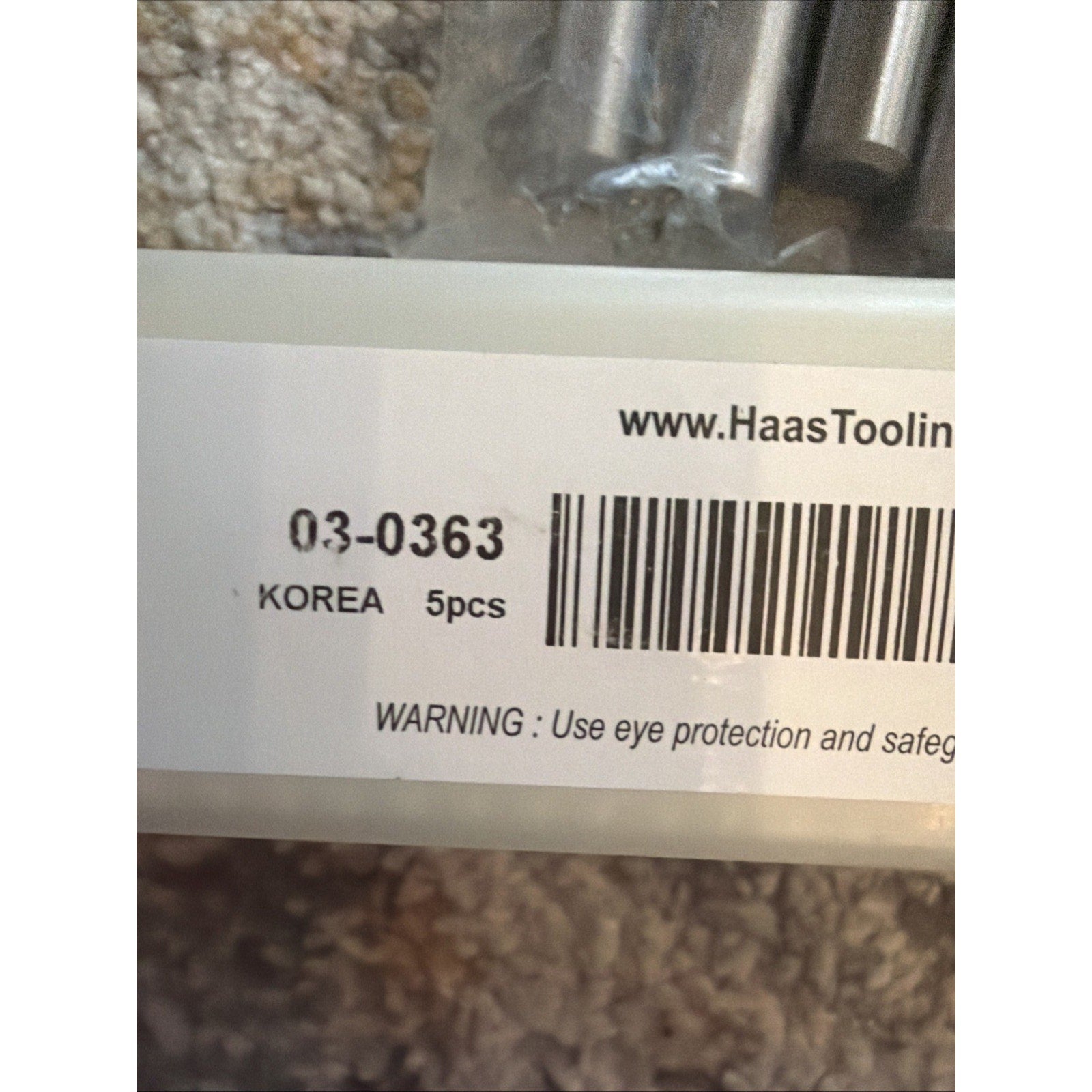 Close-up of a white label on clear plastic packaging showing a barcode, product number, origin marked as Korea, quantity of 5 pieces, and a safety warning about eye protection, placed on a beige textured surface.