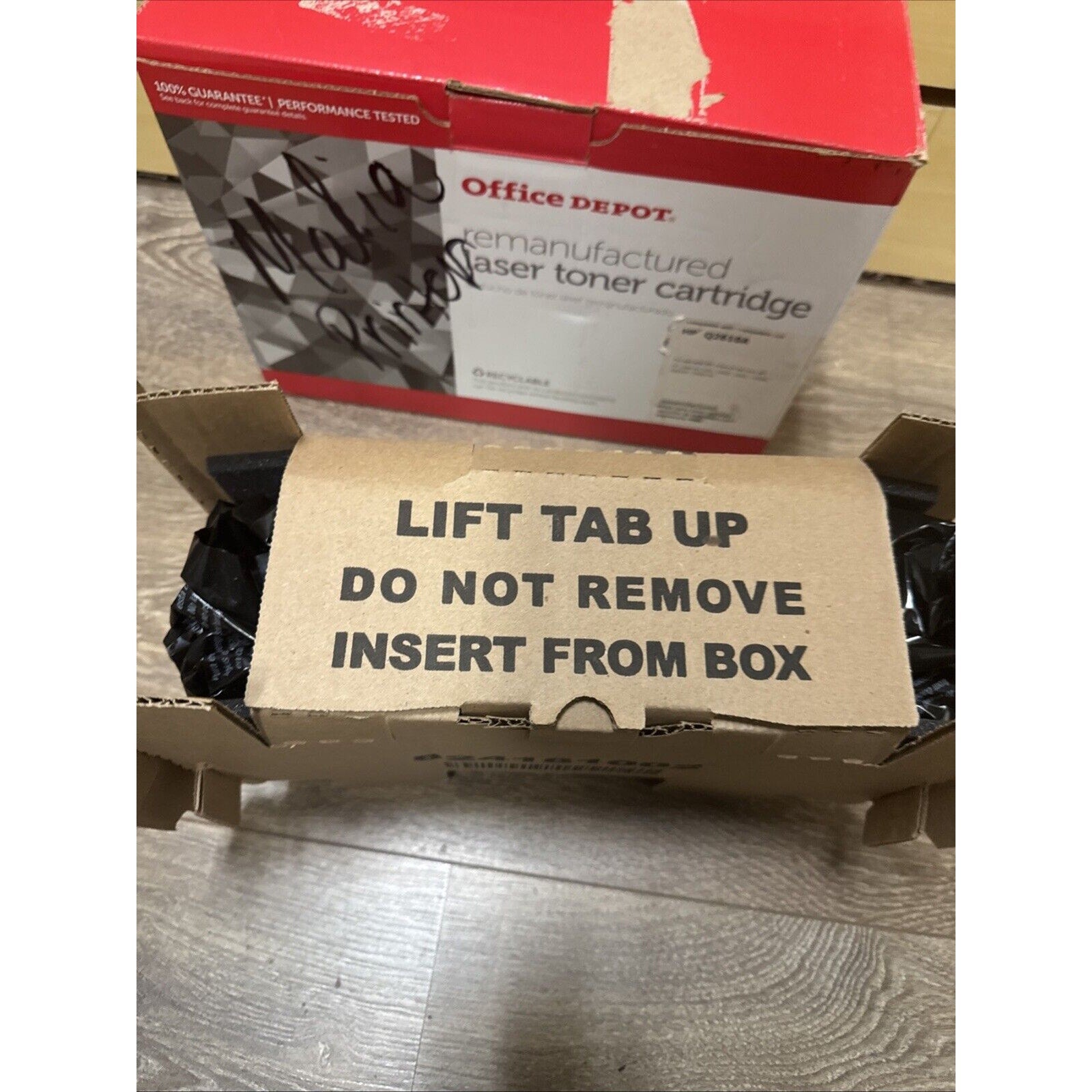 Opened cardboard box showing a brown insert with bold black text instructing "LIFT TAB UP DO NOT REMOVE INSERT FROM BOX," with a red and white Office Depot remanufactured laser toner cartridge box in the background.
