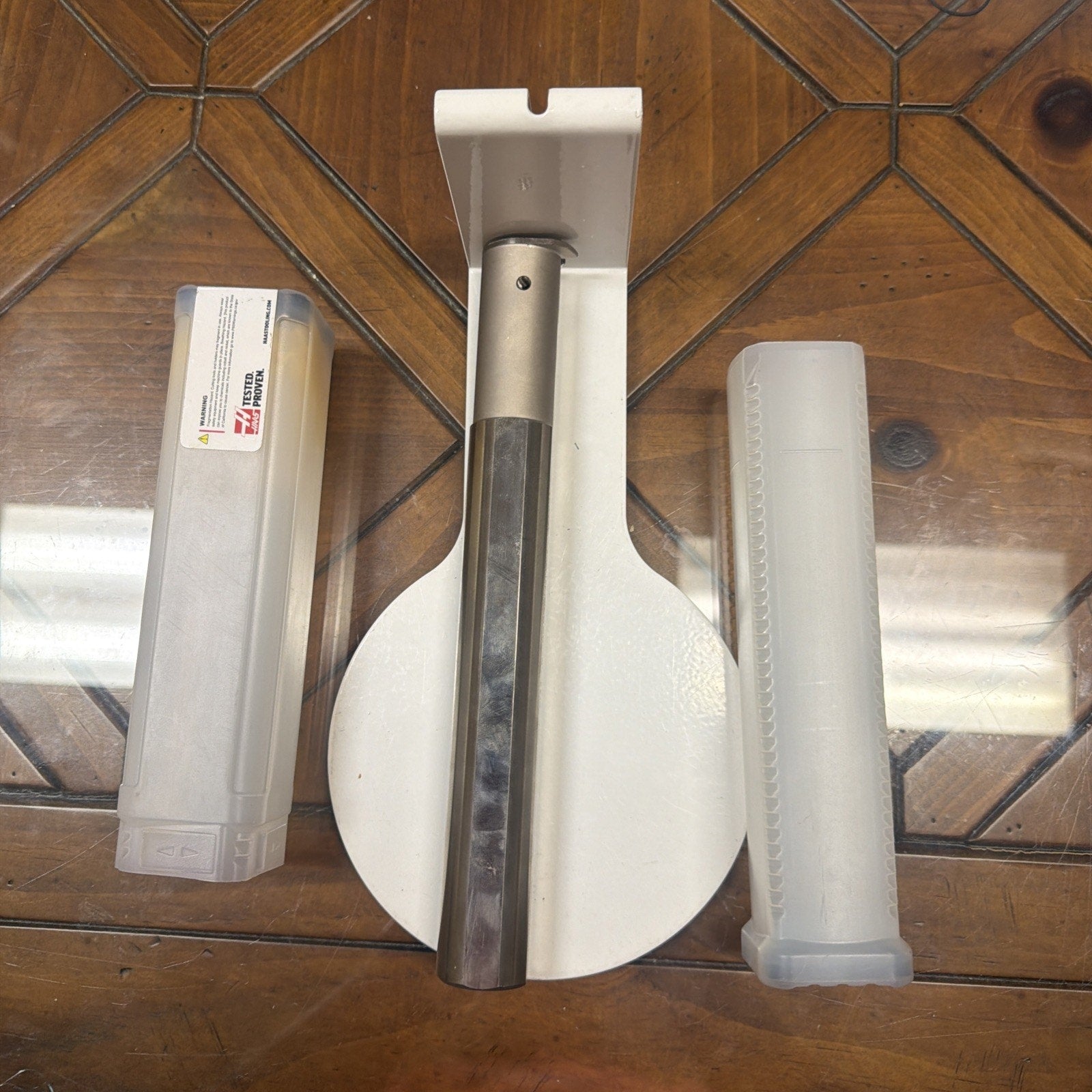Metal lathe cutting tool with integrated cylindrical end, displayed on a white holder alongside two translucent plastic protective cases on a wooden surface.