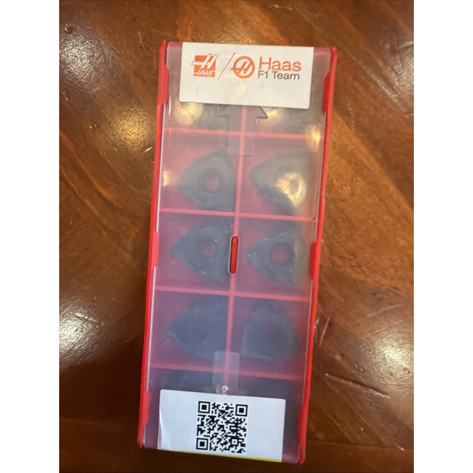 Red plastic container with a translucent lid holding six triangular metal inserts, labeled with the Haas F1 Team logo and a QR code, placed on a wooden surface.