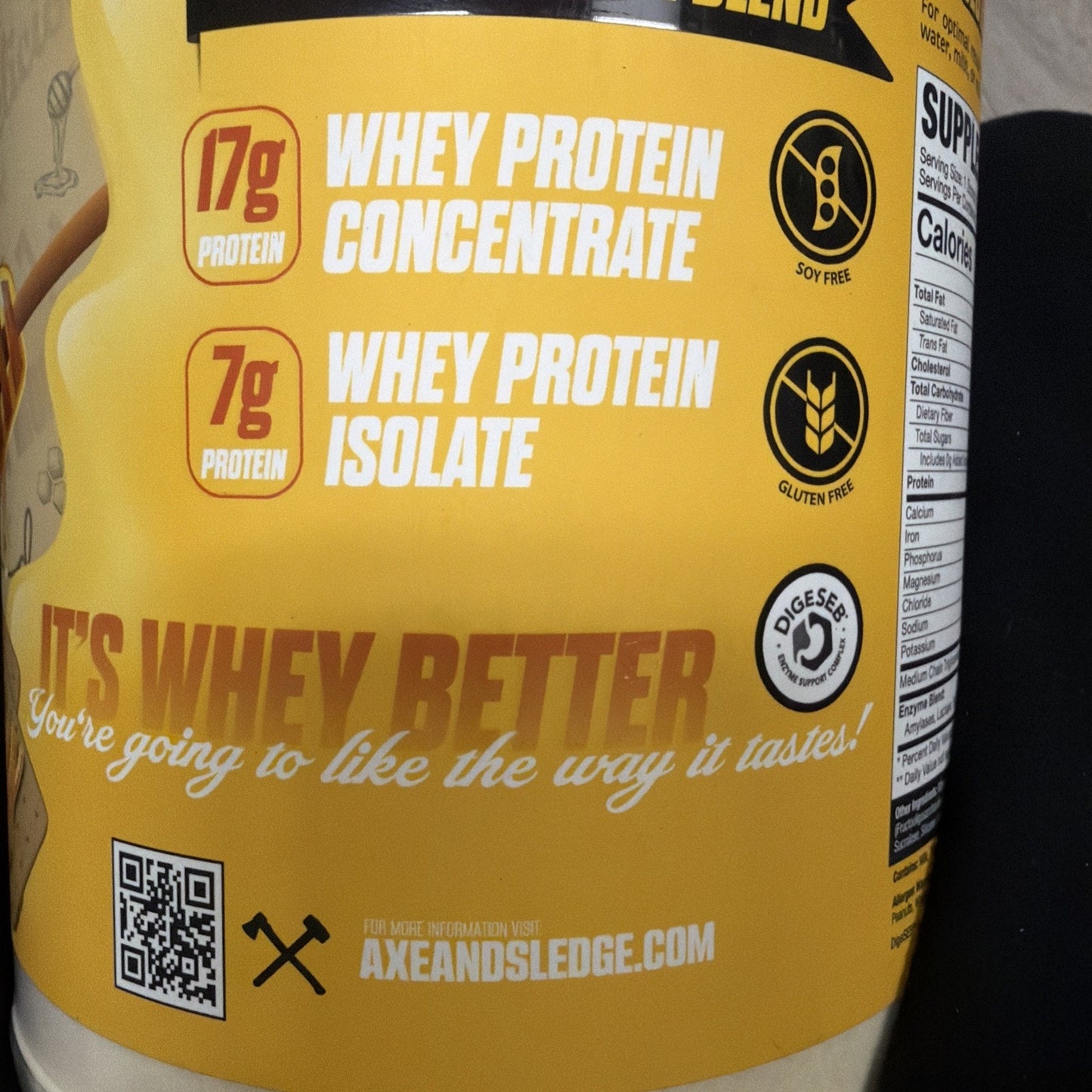 Close-up of a yellow supplement container label highlighting 17 grams of whey protein concentrate, 7 grams of whey protein isolate, soy-free and gluten-free icons, a QR code, and the slogan "It's whey better, you're going to like the way it tastes.