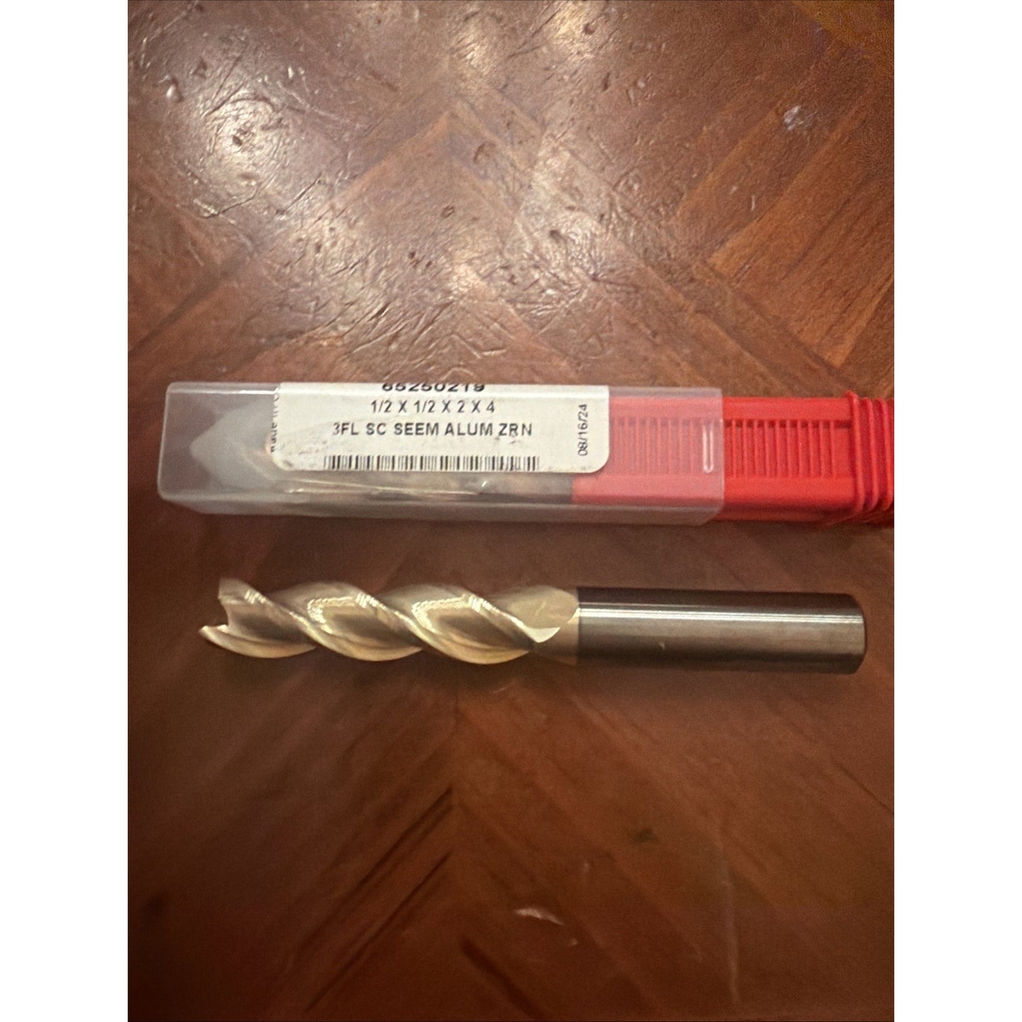Solid carbide three-flute drill bit with smooth shank, featuring spiral cutting edges, placed next to a translucent plastic case with a white label and red base on a wooden surface.