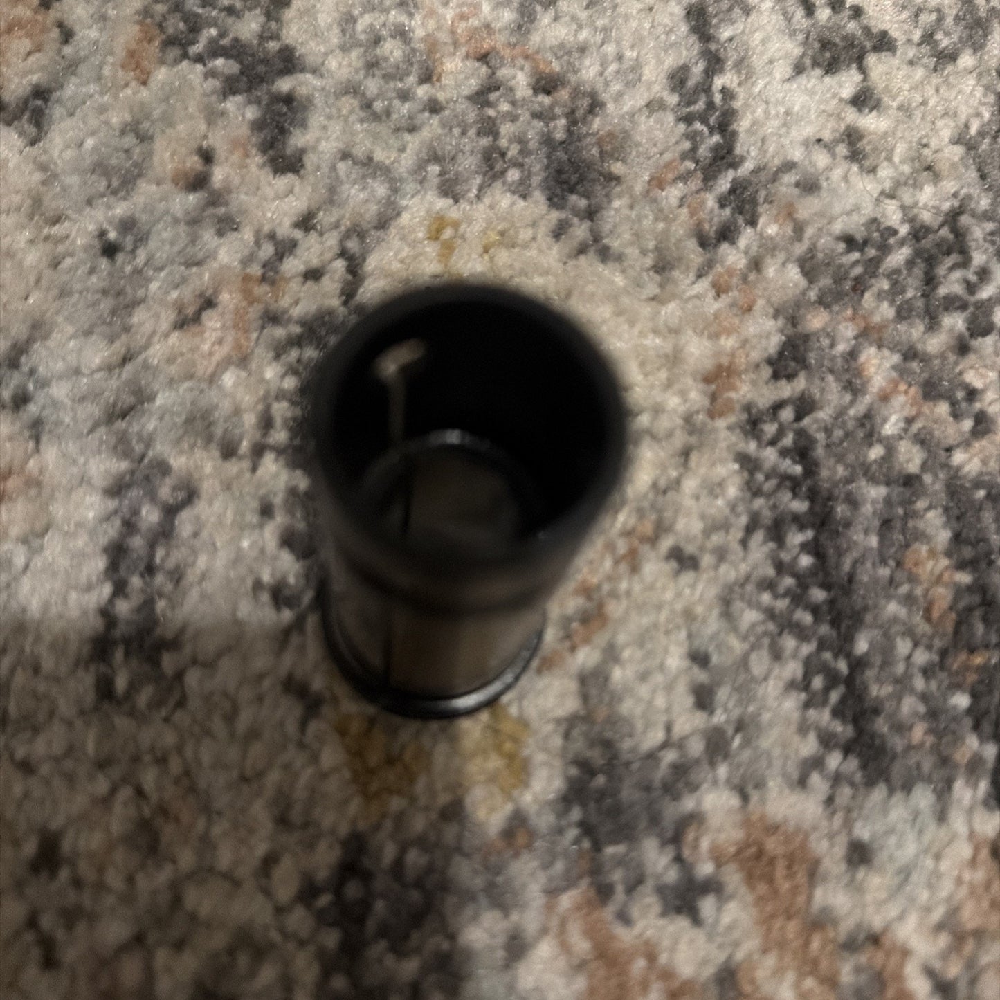 Top-down view of a small black cylindrical object standing on a textured carpet with a pattern of gray, beige, and brown hues.