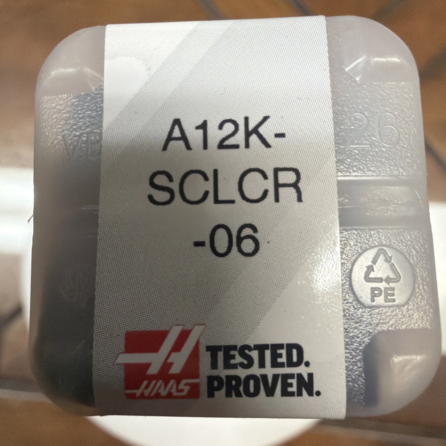 Small translucent plastic container with a white label showing the text "A12K-SCLCR-06" and a red and black "Tested. Proven." logo, placed on a brown surface.