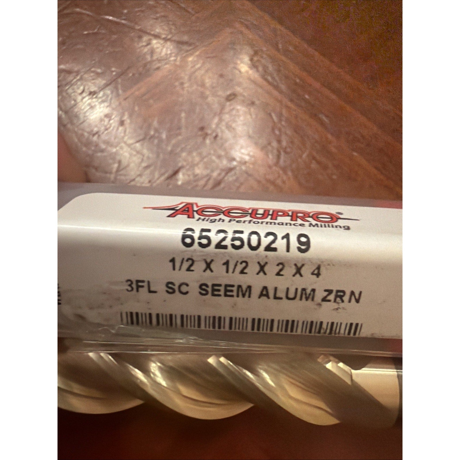 Close-up of a milling tool label showing specifications including size 1/2 x 1/2 x 2 x 4 and the text "3FL SC SEEM ALUM ZRN," with part of the spiral-fluted tool visible below.