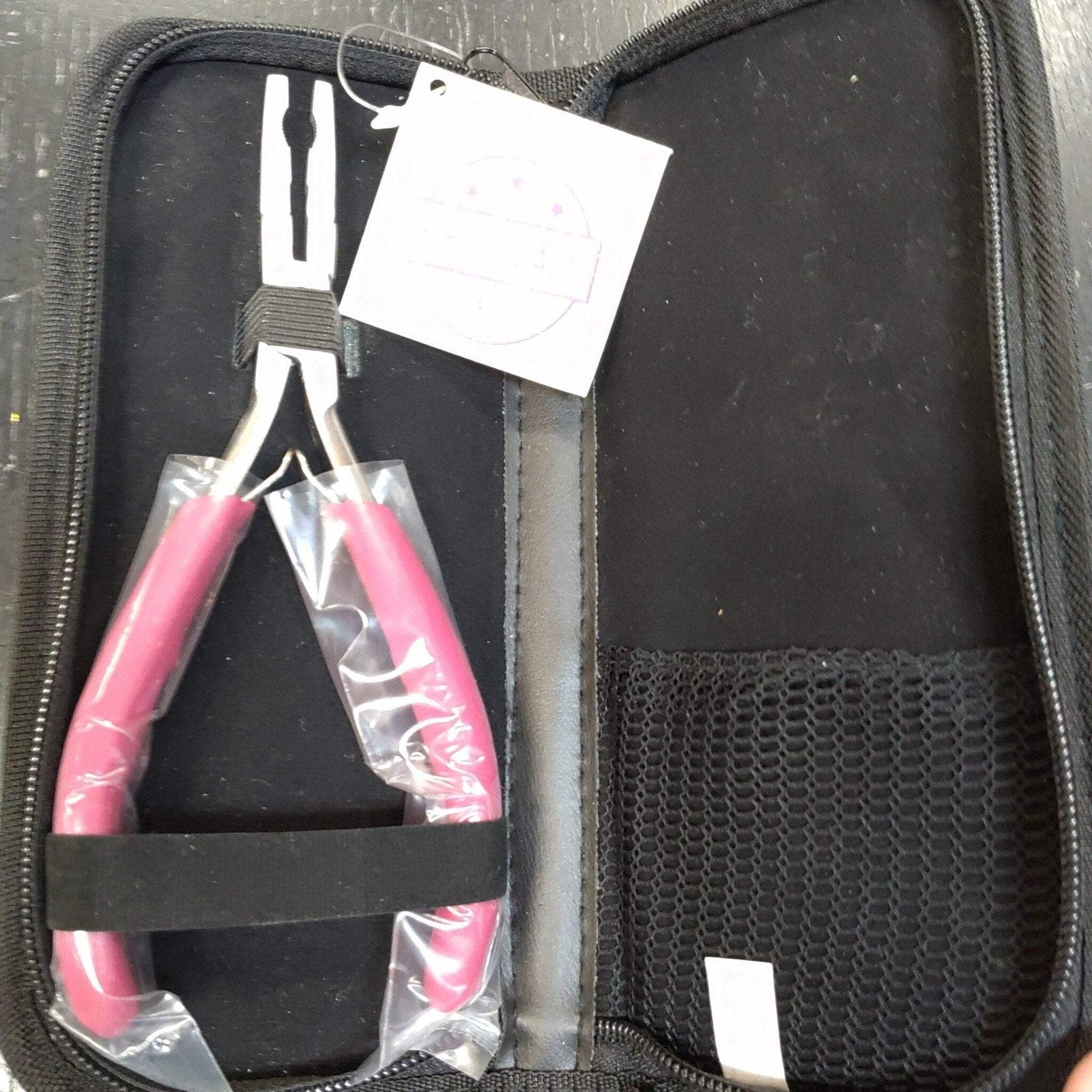 Silver pliers with bright pink handles partially wrapped in plastic, secured with a black elastic band inside an open black zippered carrying case with a mesh pocket on the right side.