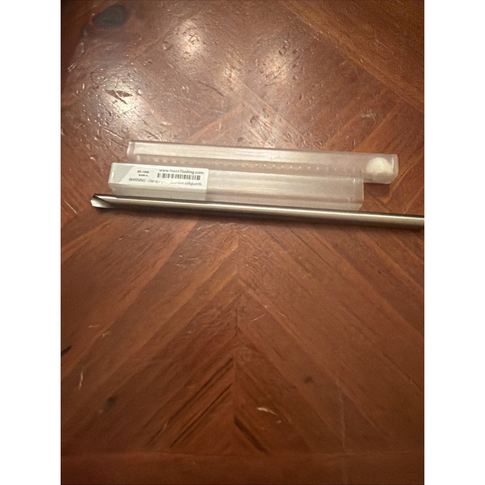 Long metal tool with a pointed tip resting on a wooden surface alongside a clear plastic protective case with a barcode label and printed text.