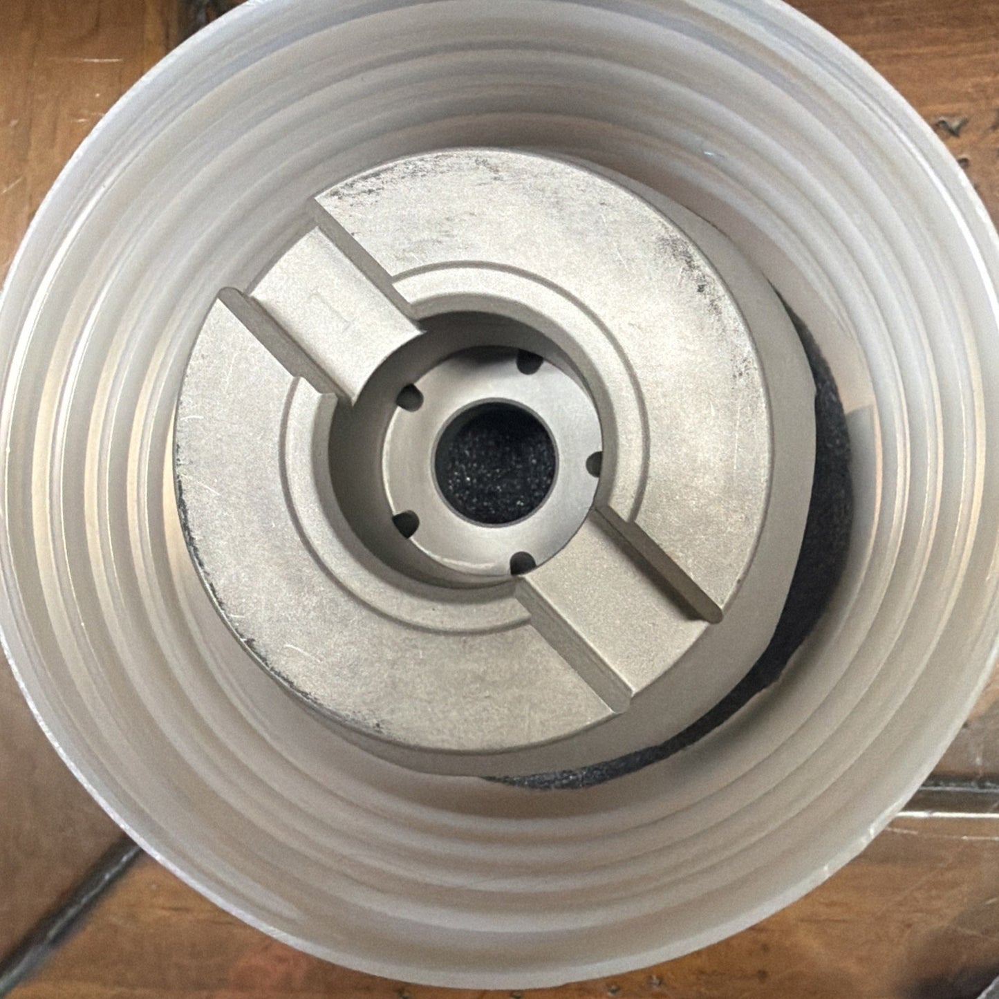 Top-down view of a cylindrical metal component with machined grooves and central holes, resting inside a white translucent plastic container on a wooden surface.
