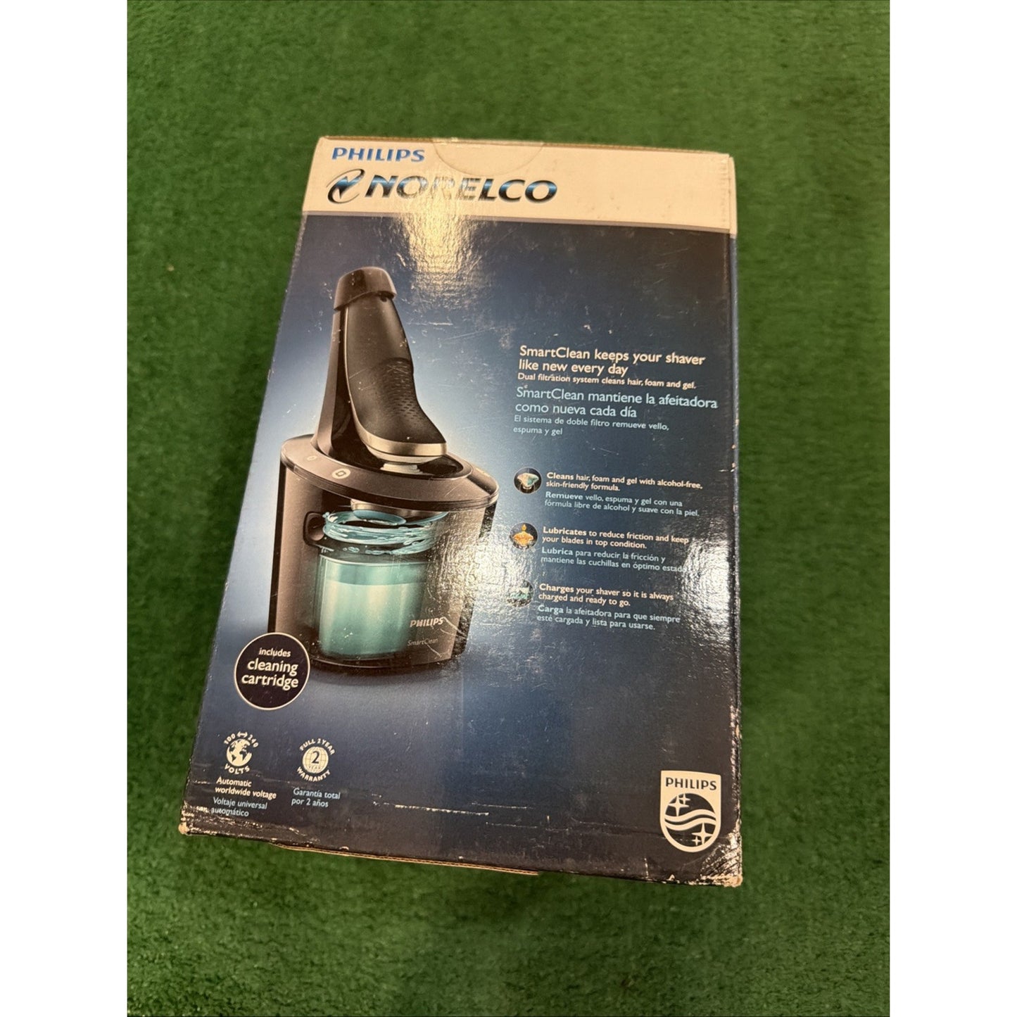 Philips Norelco electric shaver cleaning system box on a green carpet, featuring an image of the black shaver resting on the SmartClean cleaning cartridge with product details in English and Spanish.