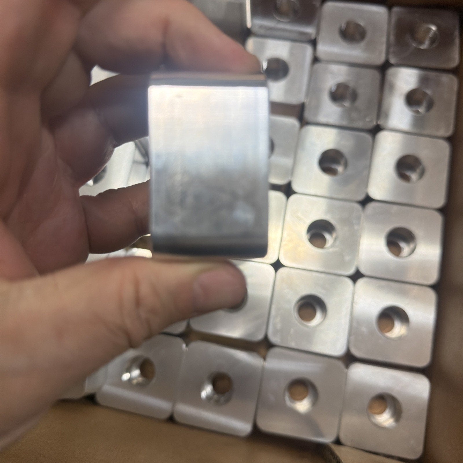 Hand holding a polished metal square block with rounded edges, in front of a box filled with similar metal blocks each featuring a centered circular hole.