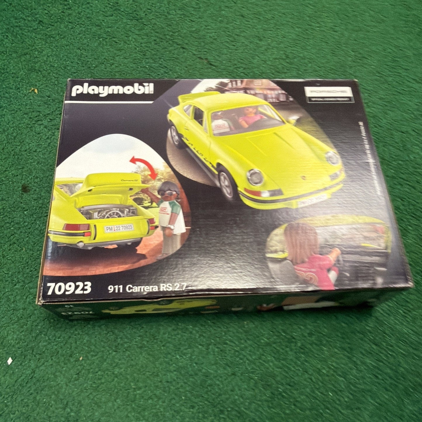 Boxed Playmobil set featuring a detailed yellow 911 Carrera RS 2.7 model car with opening trunk and driver figure holding a helmet, displayed on a green surface.