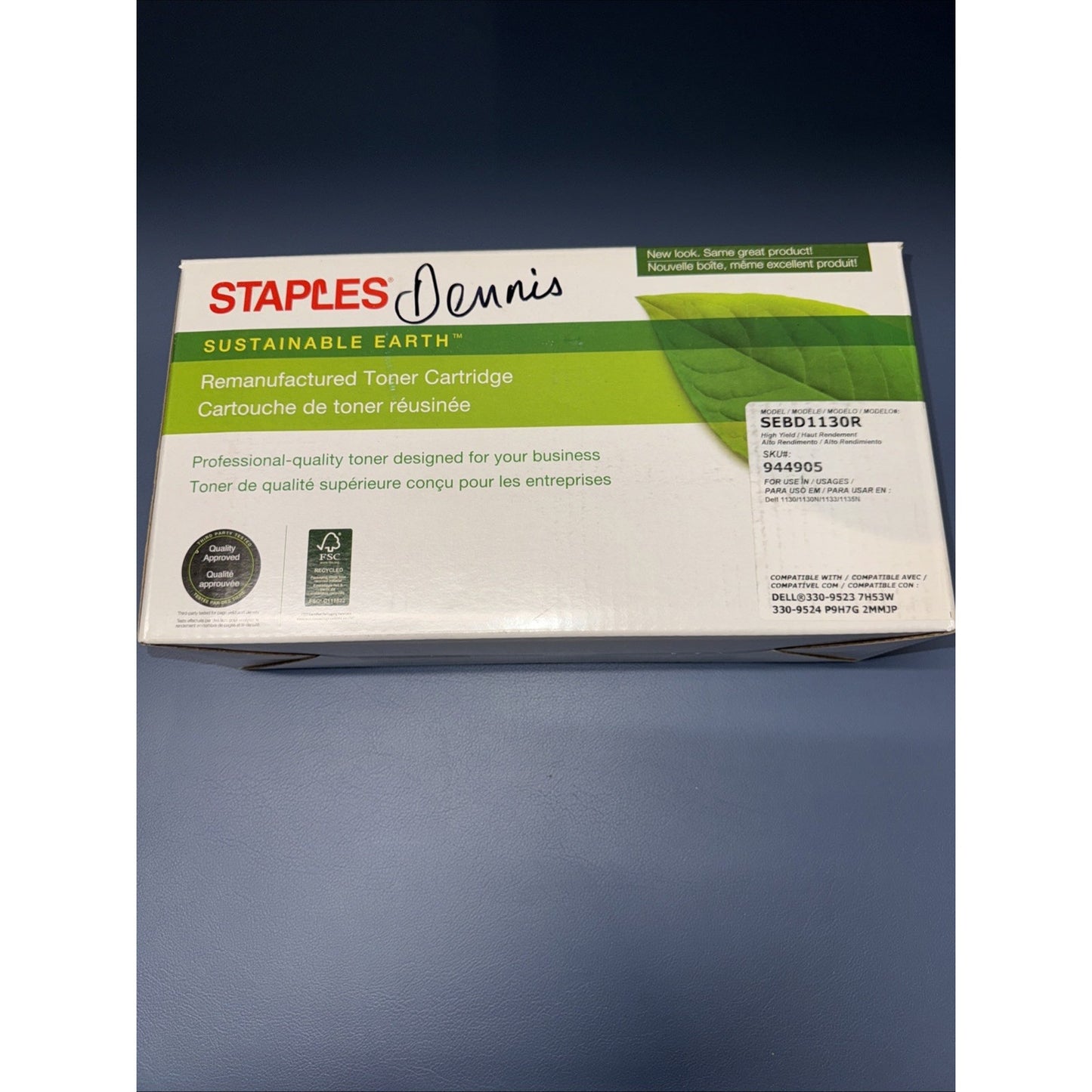 White and green packaging box for a remanufactured toner cartridge from Staples Sustainable Earth, featuring product details, a certification seal, and a handwritten name "Dennis" on the top.