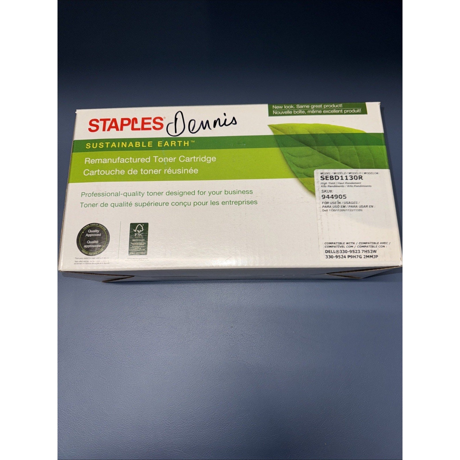 White and green packaging box for a remanufactured toner cartridge from Staples Sustainable Earth, featuring product details, a certification seal, and a handwritten name "Dennis" on the top.