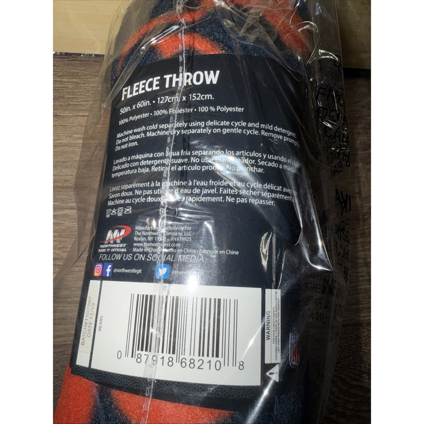 Fleece throw blanket in orange and dark blue tones, wrapped in clear plastic with a black label detailing care instructions, fabric composition, and a barcode on a wooden surface.