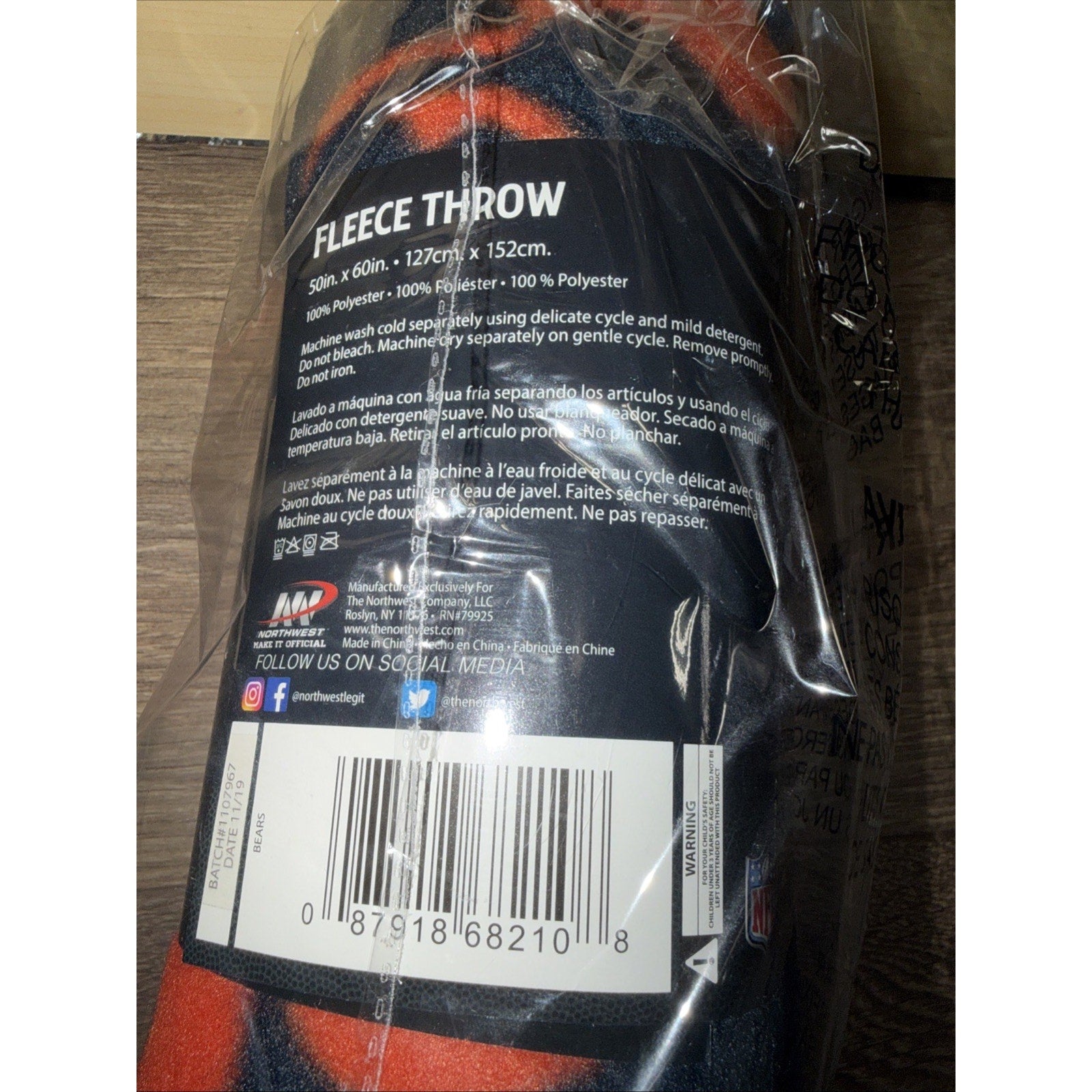 Fleece throw blanket in orange and dark blue tones, wrapped in clear plastic with a black label detailing care instructions, fabric composition, and a barcode on a wooden surface.