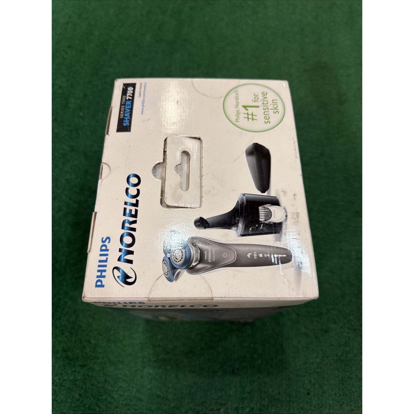 White retail box for a Philips Norelco electric shaver showing the shaver, nose trimmer, and protective cap, placed on a green textured surface with branding and product claims.