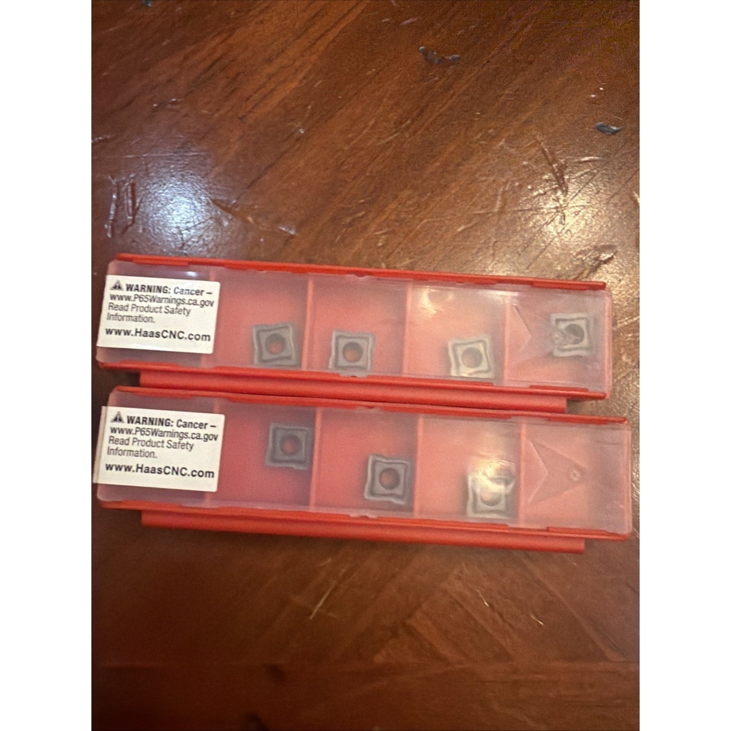 Two transparent red plastic trays containing small square metal inserts, each compartment holding one insert, resting on a wooden surface with warning labels attached to the trays.
