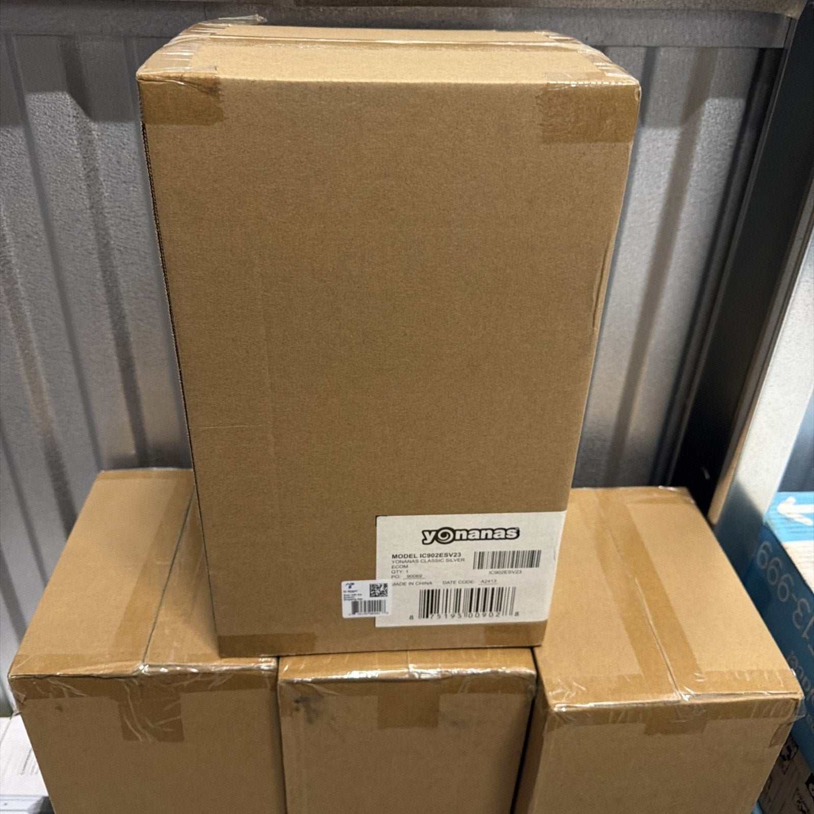 Stack of four brown cardboard shipping boxes sealed with tape, one box on top featuring a white label with the brand name "Yonanas" and product information, placed in a metallic storage environment.