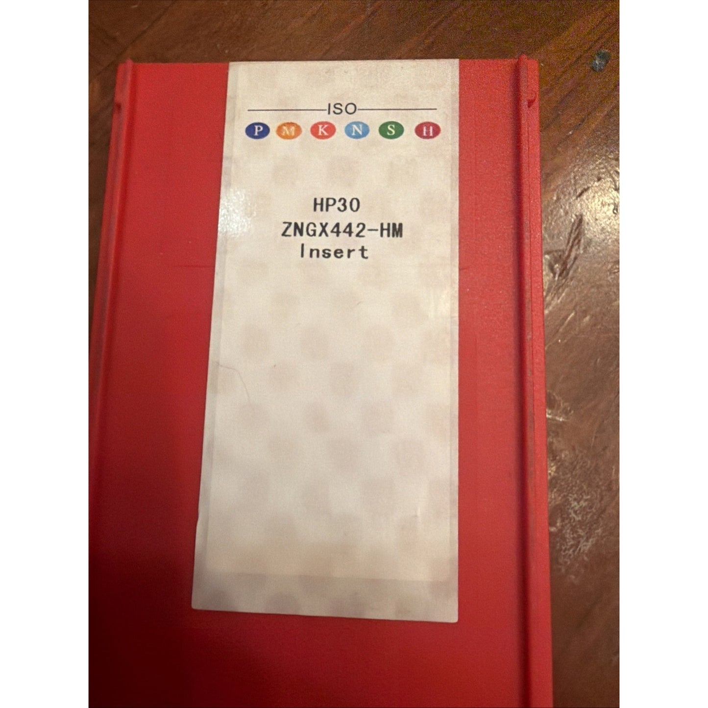 Red rectangular plastic container with a white label displaying "HP30 ZNGX442-HM Insert" and colored ISO classification symbols, placed on a wooden surface.