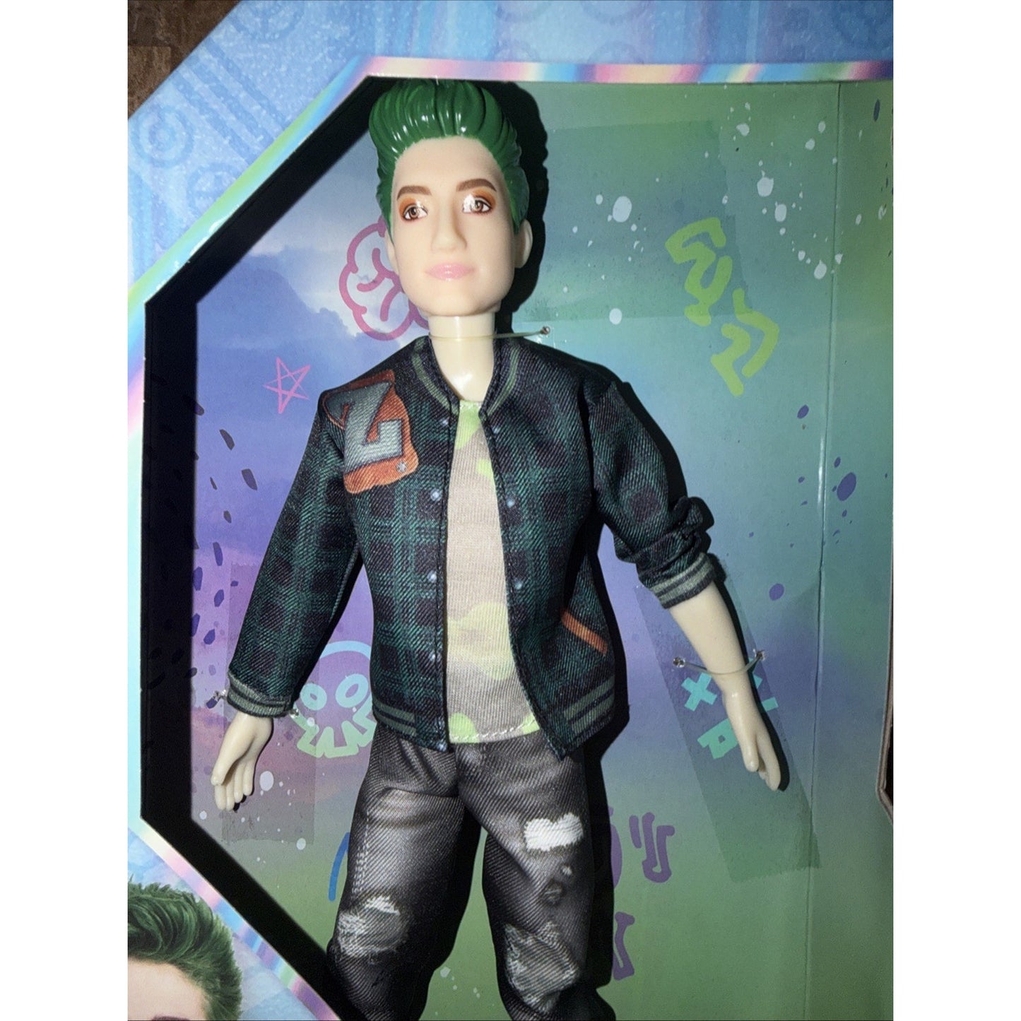 Poseable doll with green hair wearing a green and black patterned jacket over a graphic t-shirt and black distressed pants, displayed inside a colorful illustrated box.