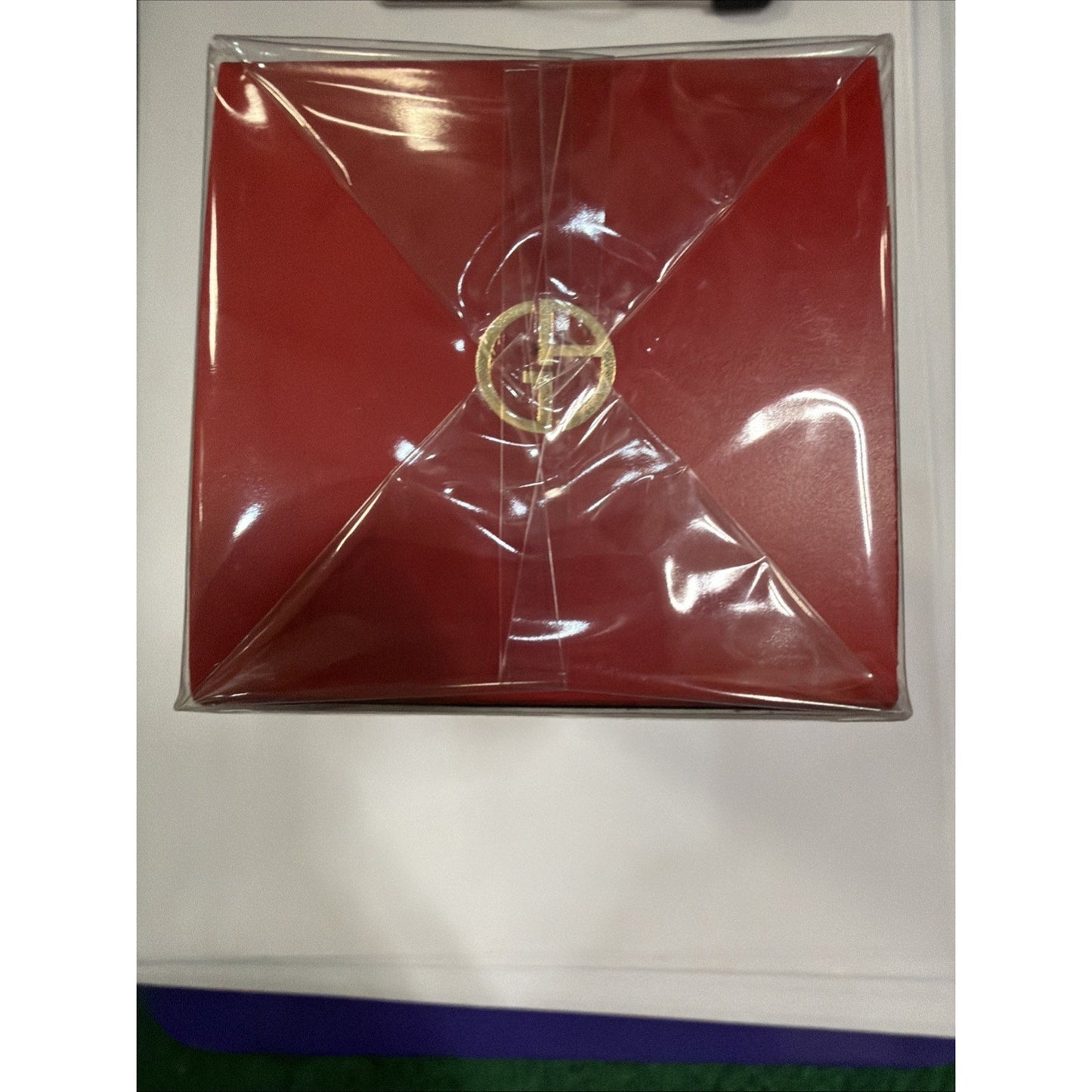 Top view of a red square box wrapped with clear plastic film sealed at the center, resting on a white surface with a hint of purple and green edges underneath.